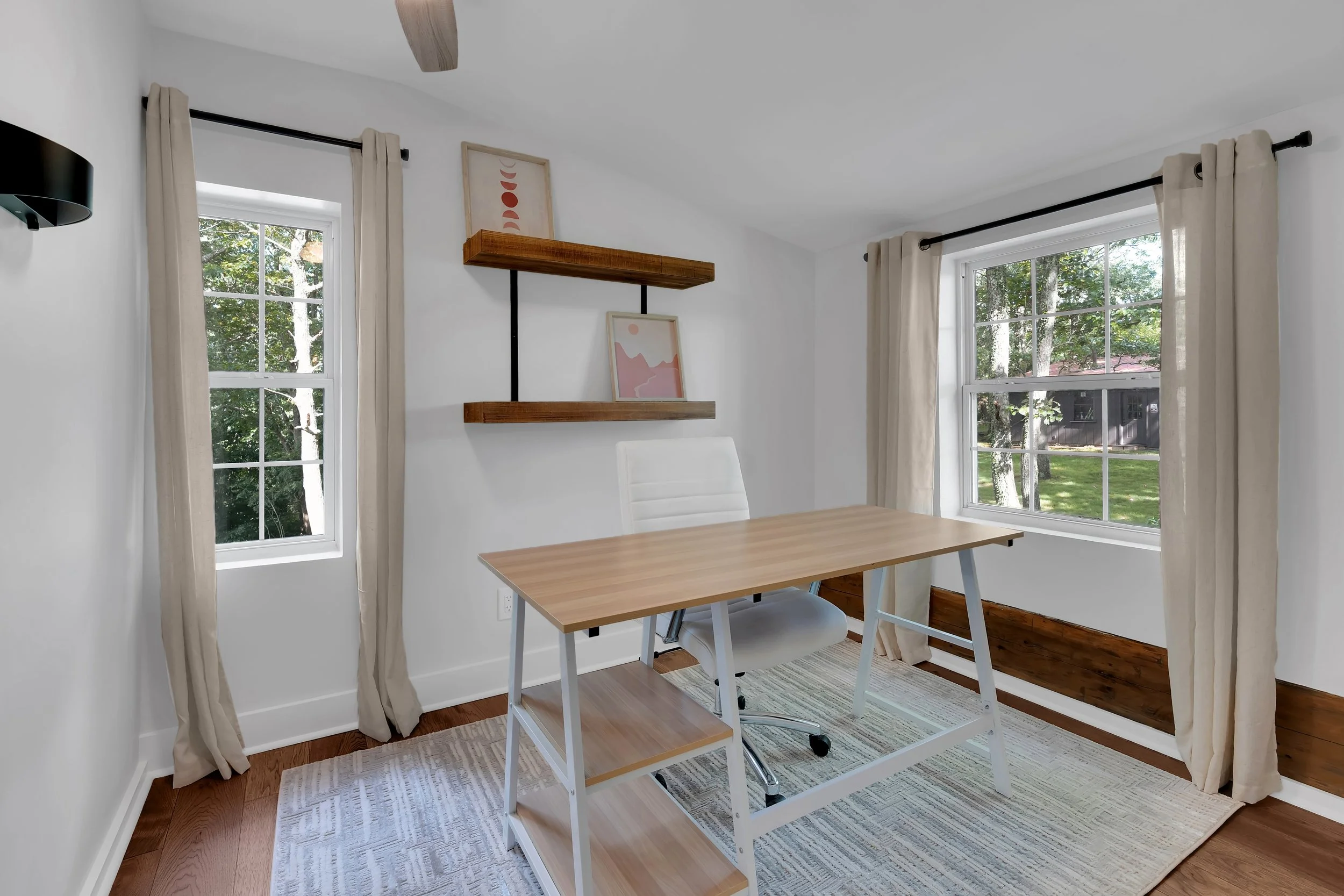 Home office with a wooden desk, white chair, two windows with beige curtains, and wall shelves with framed art, in a bright room with hardwood floors and a neutral rug.