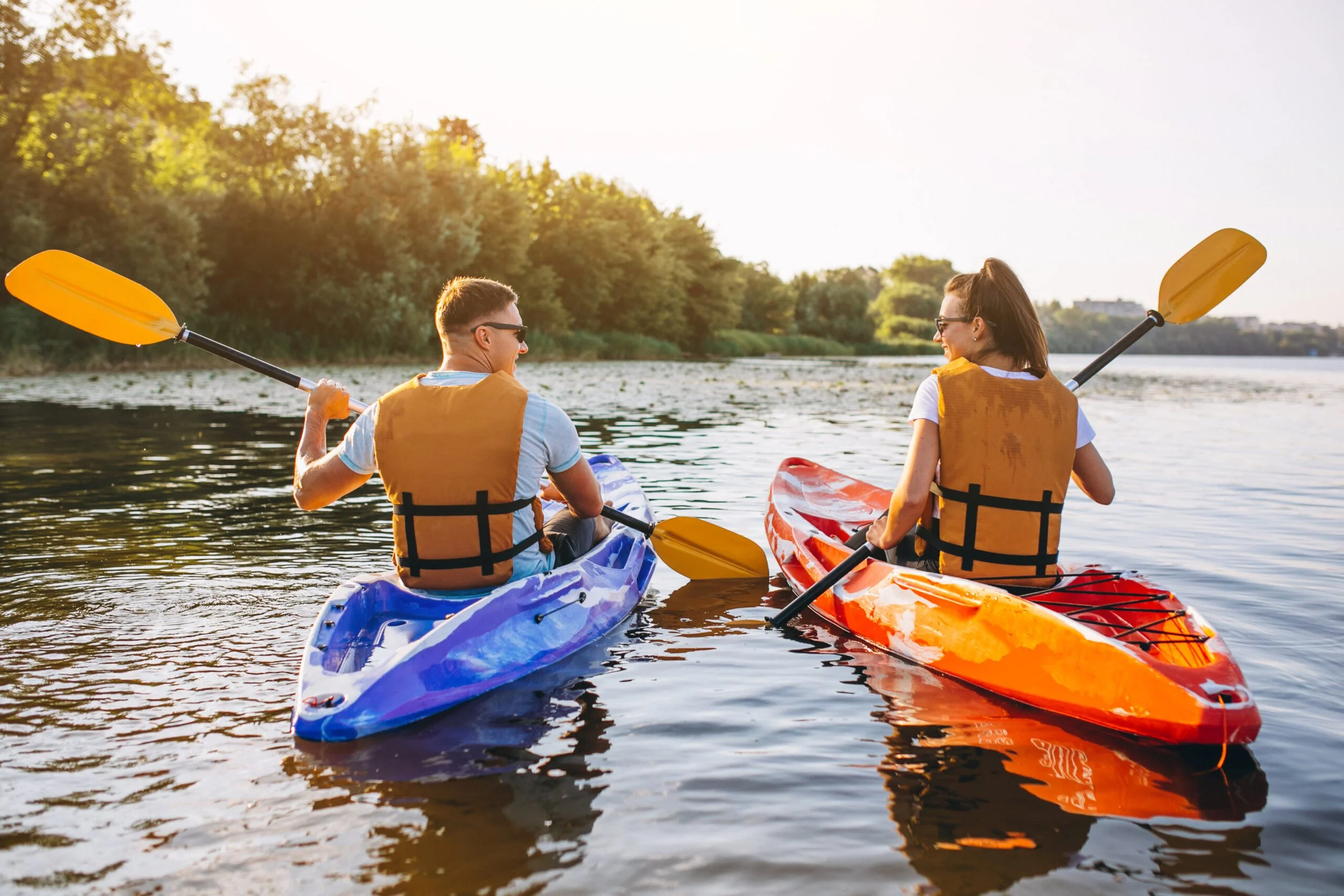 Two people kayaking on a calm river during sunset, wearing life jackets and sunglasses, with lush trees in the background.