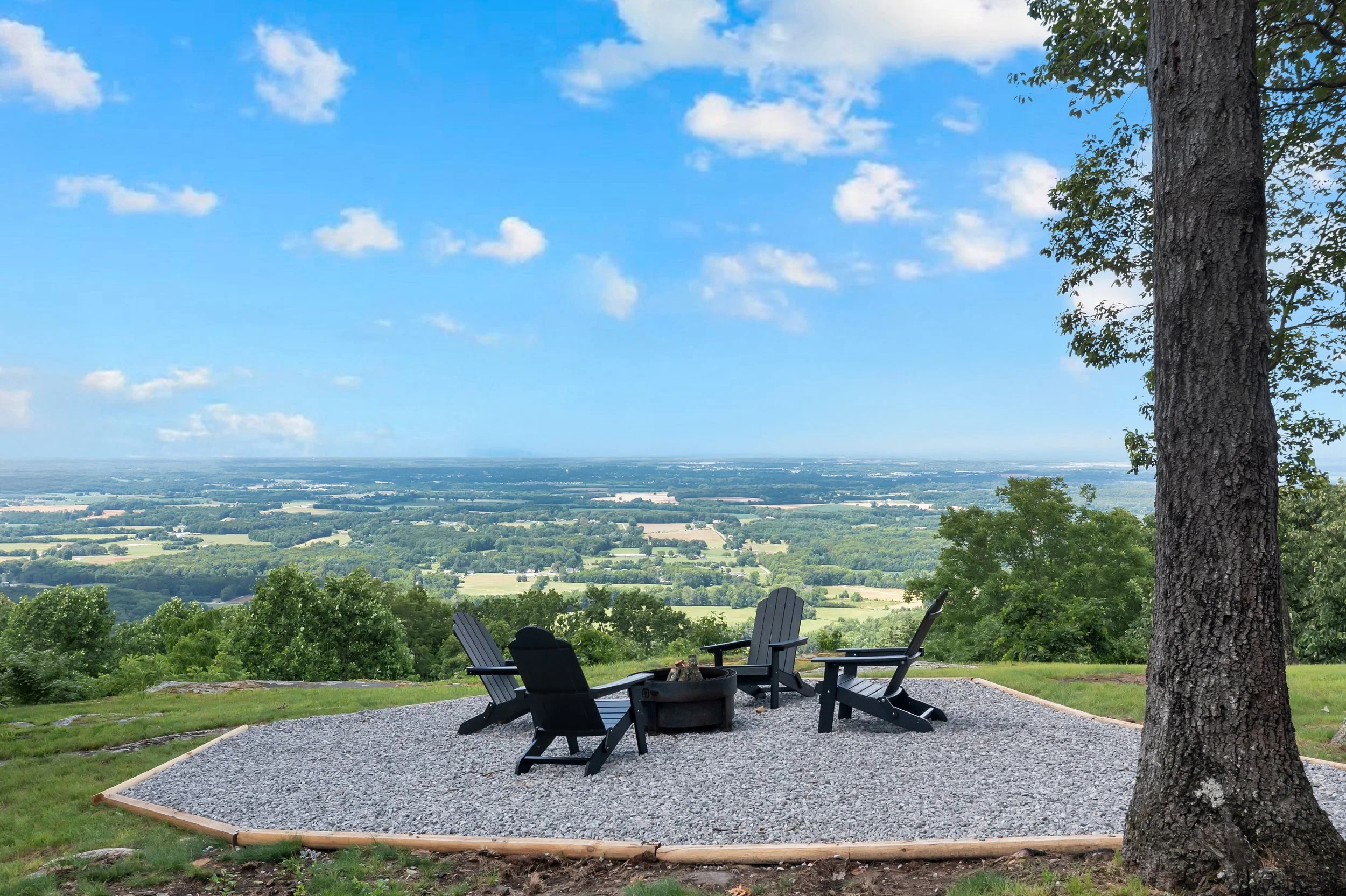 A scenic outdoor sitting area with four black Adirondack chairs arranged around a fire pit on a gravel pad. There are tall trees and a wide view of lush green fields and a blue sky with scattered clouds in the background. Gorgeous views.