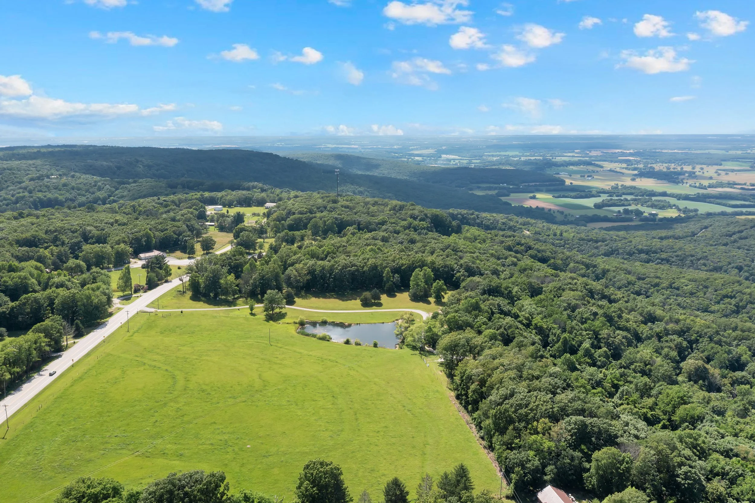 Aerial view of green rolling hills, dense forests, a small pond, and a rural road under a partly cloudy blue sky.