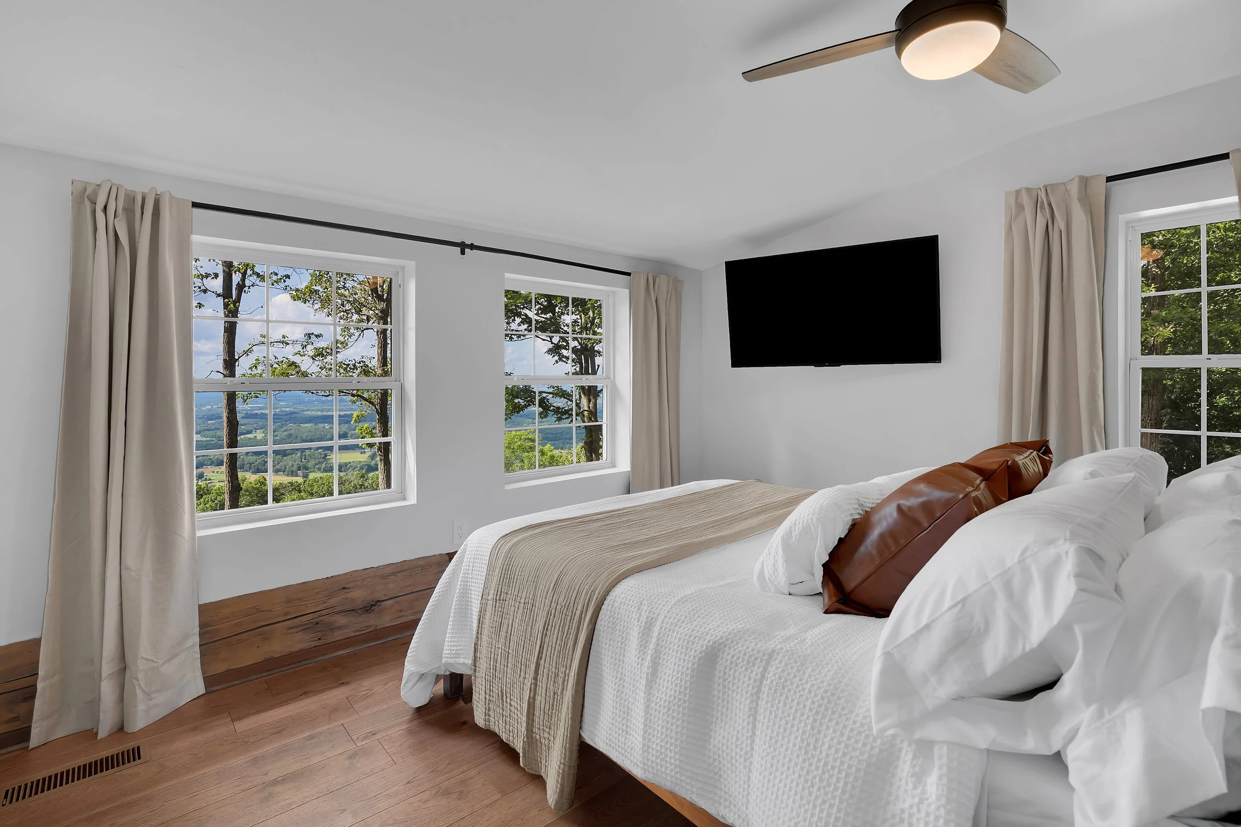 Bright bedroom with three large windows overlooking a scenic landscape, white walls, wooden floor, a bed with white bedding and brown and white pillows, beige curtains, and a wall-mounted TV.
