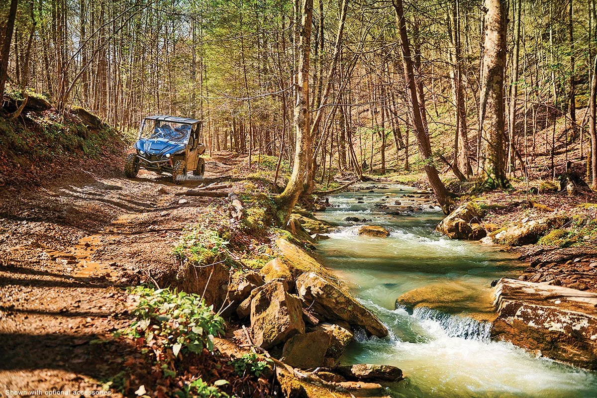 An all-terrain vehicle (ATV) riding on a dirt forest trail next to a flowing creek, surrounded by trees and rocks.