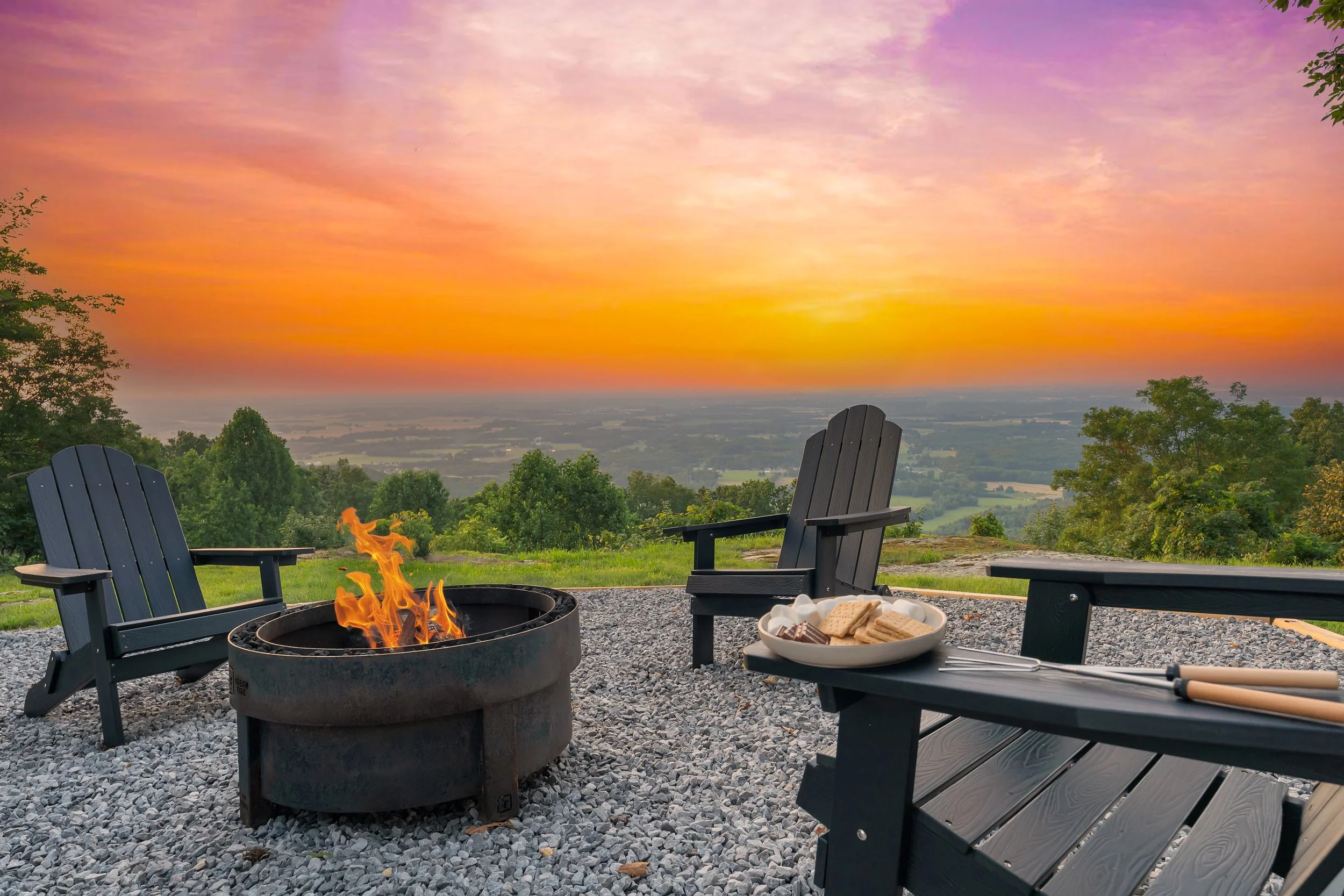 A scenic outdoor patio at sunset with three black Adirondack chairs around a fire pit, a plate of assorted cookies on one chair, and a pair of tongs resting on another chair. The sky displays orange, pink, and purple hues over a lush green landscape.