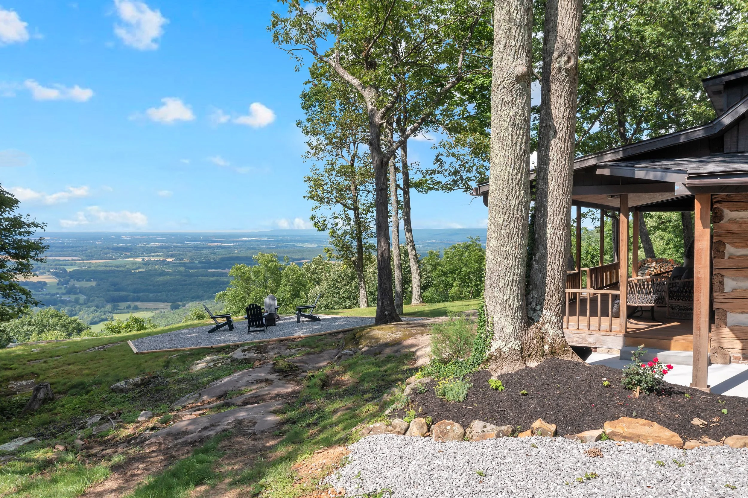 A scenic backyard overlooking a valley with a view of trees and farmland, including a small patio area with four black chairs and a fire pit, and a wooden house with a porch.