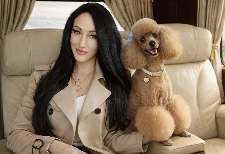 A woman with long black hair and a beige trench coat sitting on a beige couch with a Poodle dog.