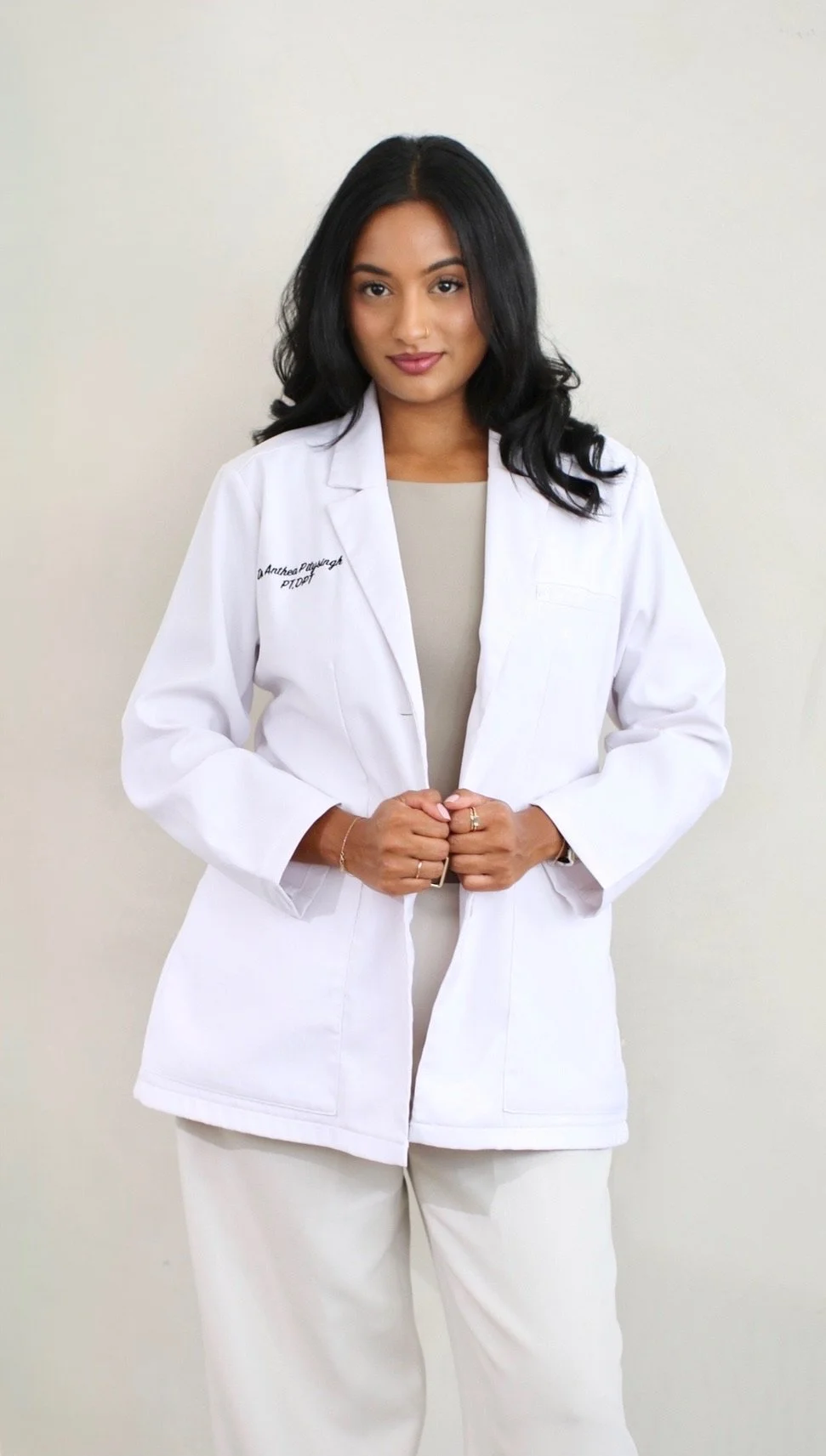 Woman in a white medical coat standing against a plain wall.