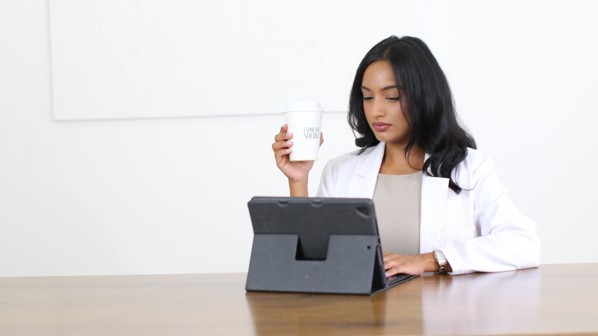A woman with dark hair, wearing a white coat, sitting at a wooden desk, holding a coffee cup in her right hand, and using a tablet with a black cover on the desk.