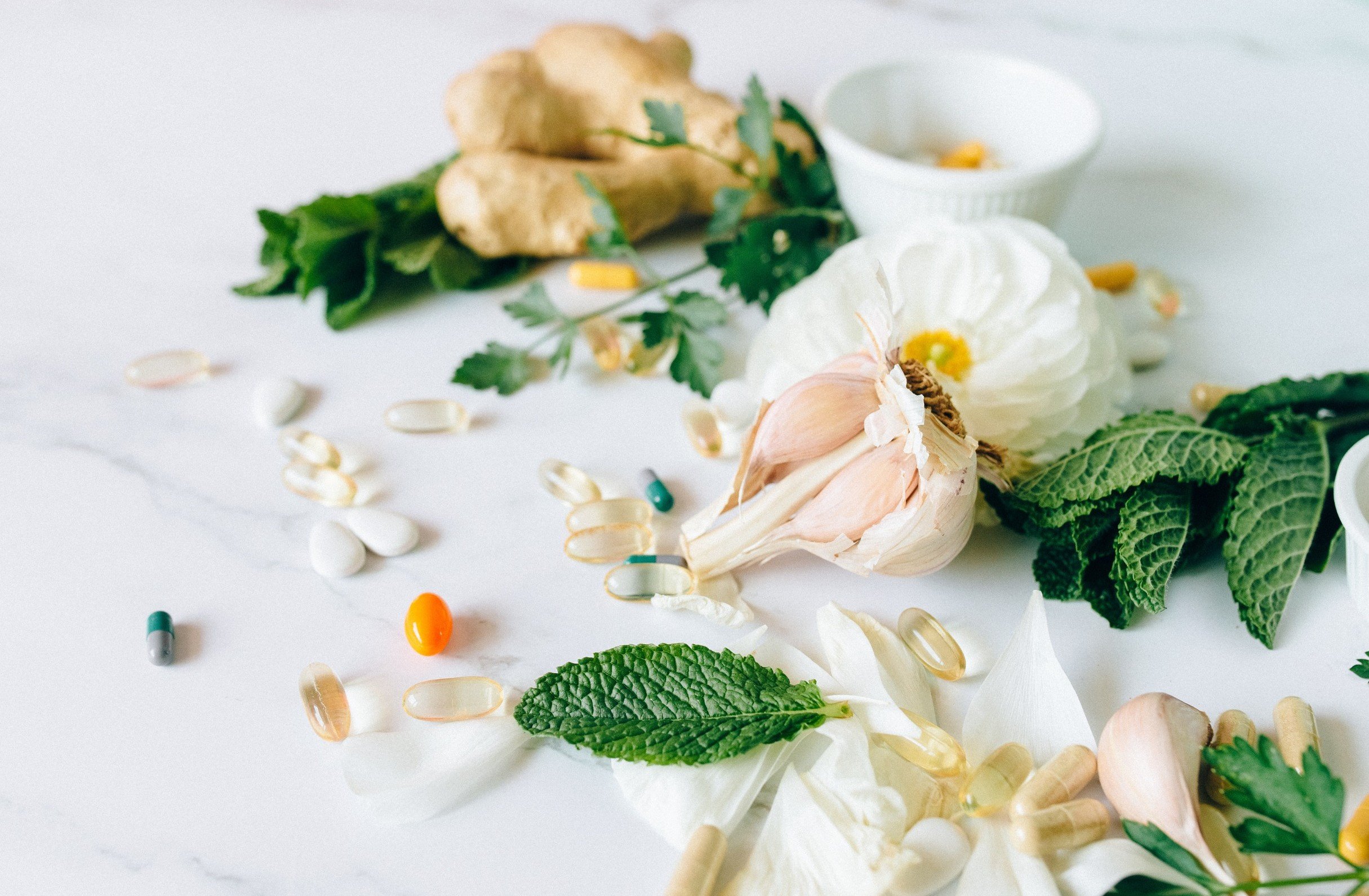 A flat lay of fresh herbs, garlic, ginger, pills, capsules, and a small bowl on a white surface.
