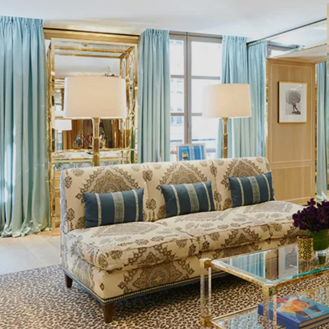 Living room with a patterned sofa, blue pillows, light blue curtains, and gold lamps with beige shades.
