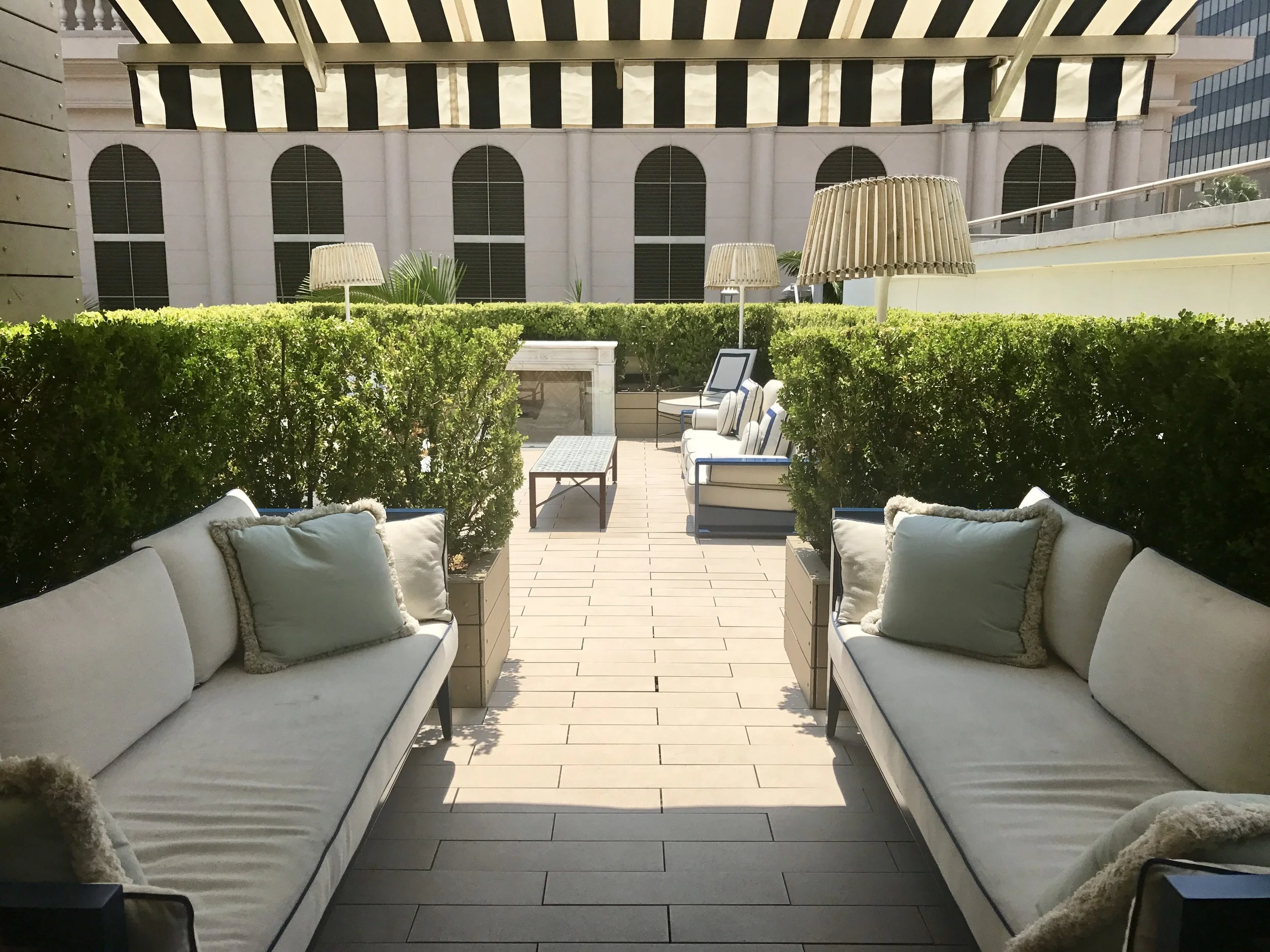 Outdoor terrace seating area with white sofas and decorative cushions, overlooking a green hedge, on a tiled terrace with large umbrellas. Blue and white striped awning 