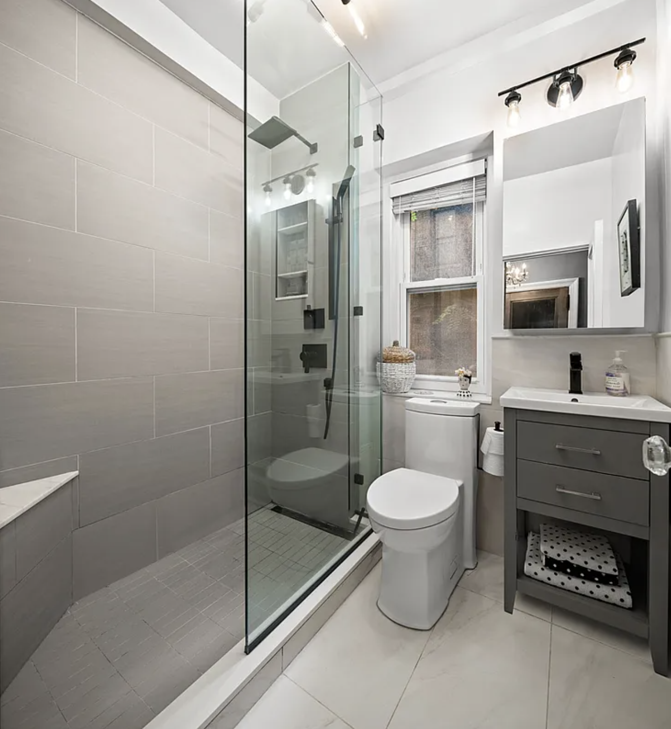 Modern bathroom with a glass-enclosed shower, a toilet, a gray vanity with a small sink, a mirror, and a window with white blinds, decorated with a woven basket and small decorative items.