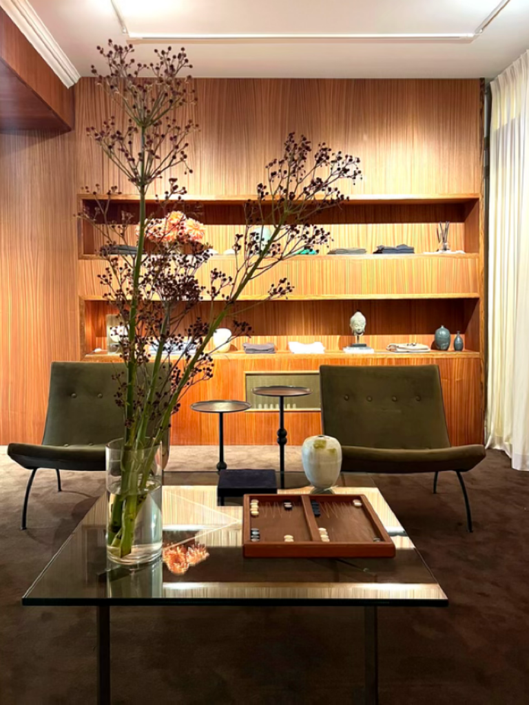 A modern living room with a wooden wall and built-in shelves, featuring two green armchairs, a glass-top coffee table with decorative items and a backgammon set, a tall vase with flowers, and curtains on the right side.