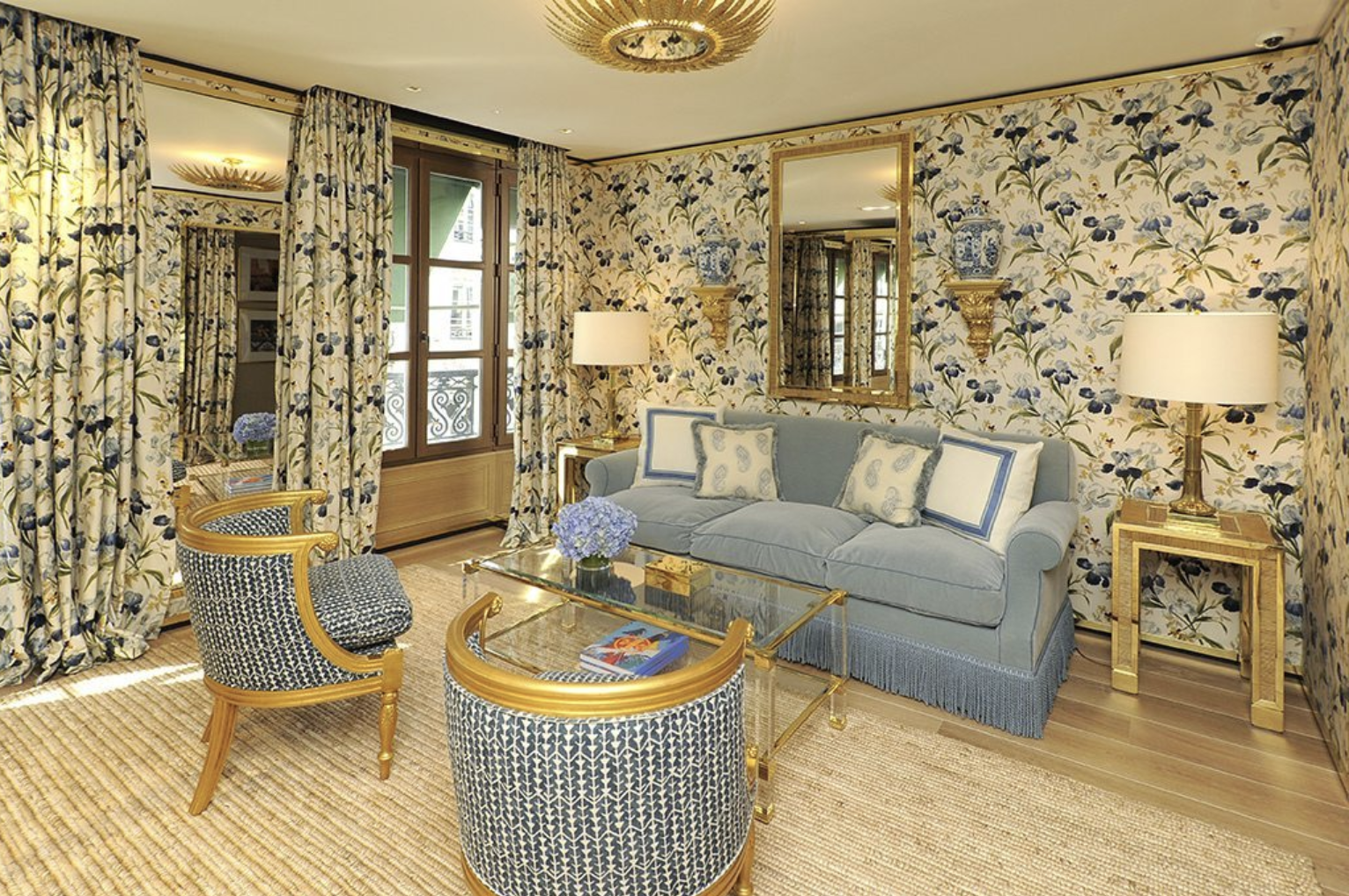 Living room with floral wallpaper, gold accents, blue sofa, armchairs, glass coffee table, lamps, mirrors, and French doors with floral curtains. Decorative prints pop through-out. 
