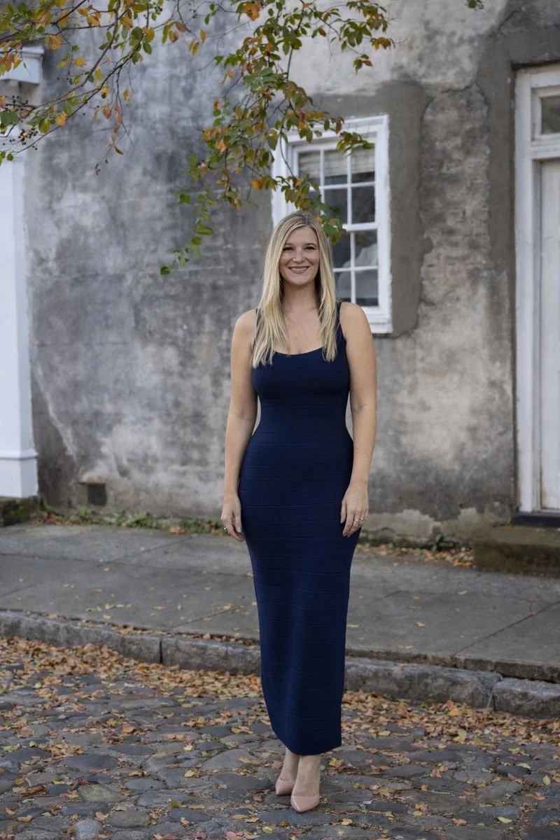 A young woman with long blond hair wearing a fitted, sleeveless navy blue dress and nude high heels, standing on a Charleston cobblestone street with fallen leaves in front of an old weathered building with a window and a white door, holding a smile