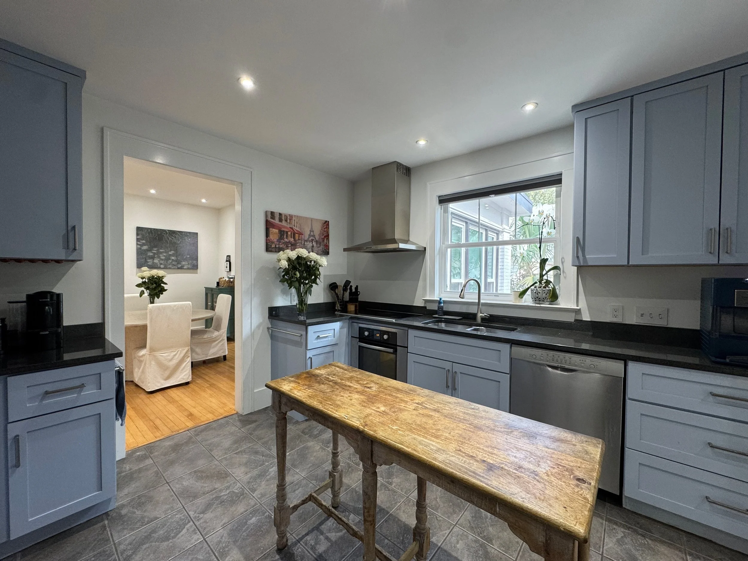 Kitchen with blue cabinets, black countertops, a wooden table, window with potted plant, and stainless steel appliances, connecting to a dining room with white chairs and table.