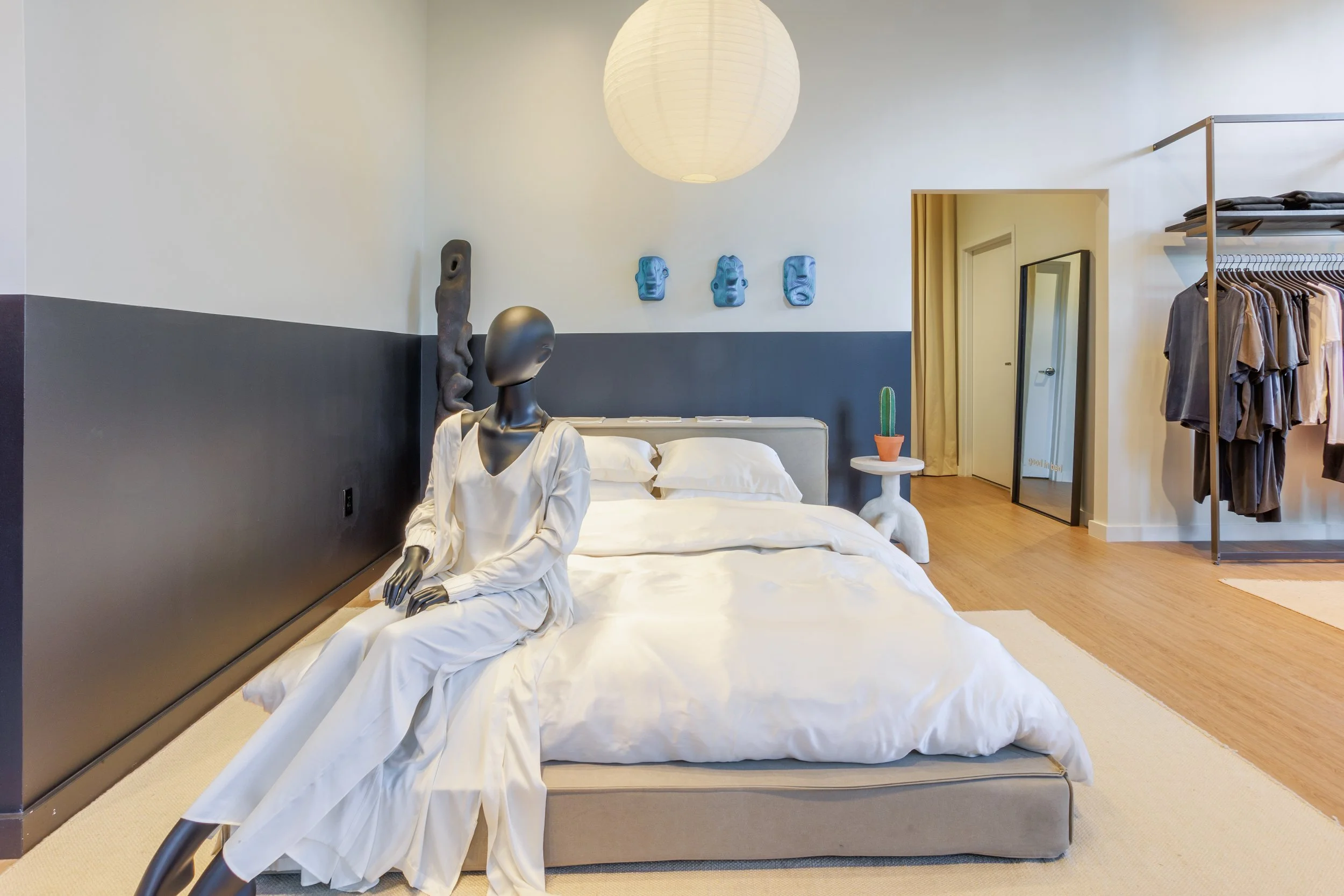 A modern bedroom featuring a mannequin sitting on a bed dressed in white, with three colorful masks on the wall above. The room has a white and dark gray color scheme, a white round paper lantern hanging from the ceiling, a cactus on a small side table, and an open clothing rack with hanging shirts.