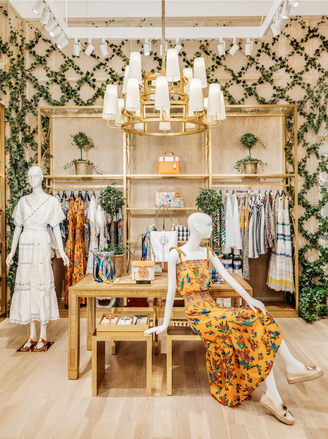 A retail clothing store with mannequins dressed in summer outfits, shelves with handbags, and racks of colorful dresses. The decor includes lattice with green plants and ivy and a wooden backdrop with a large chandelier overhead.