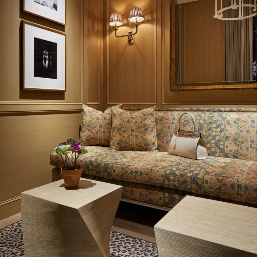 A cozy sitting area with a patterned sofa and matching pillows, a small coffee table with a potted plant, a handbag on the sofa, framed black and white photographs on the wall, a wall-mounted lamp, and a mirror reflecting part of the room.