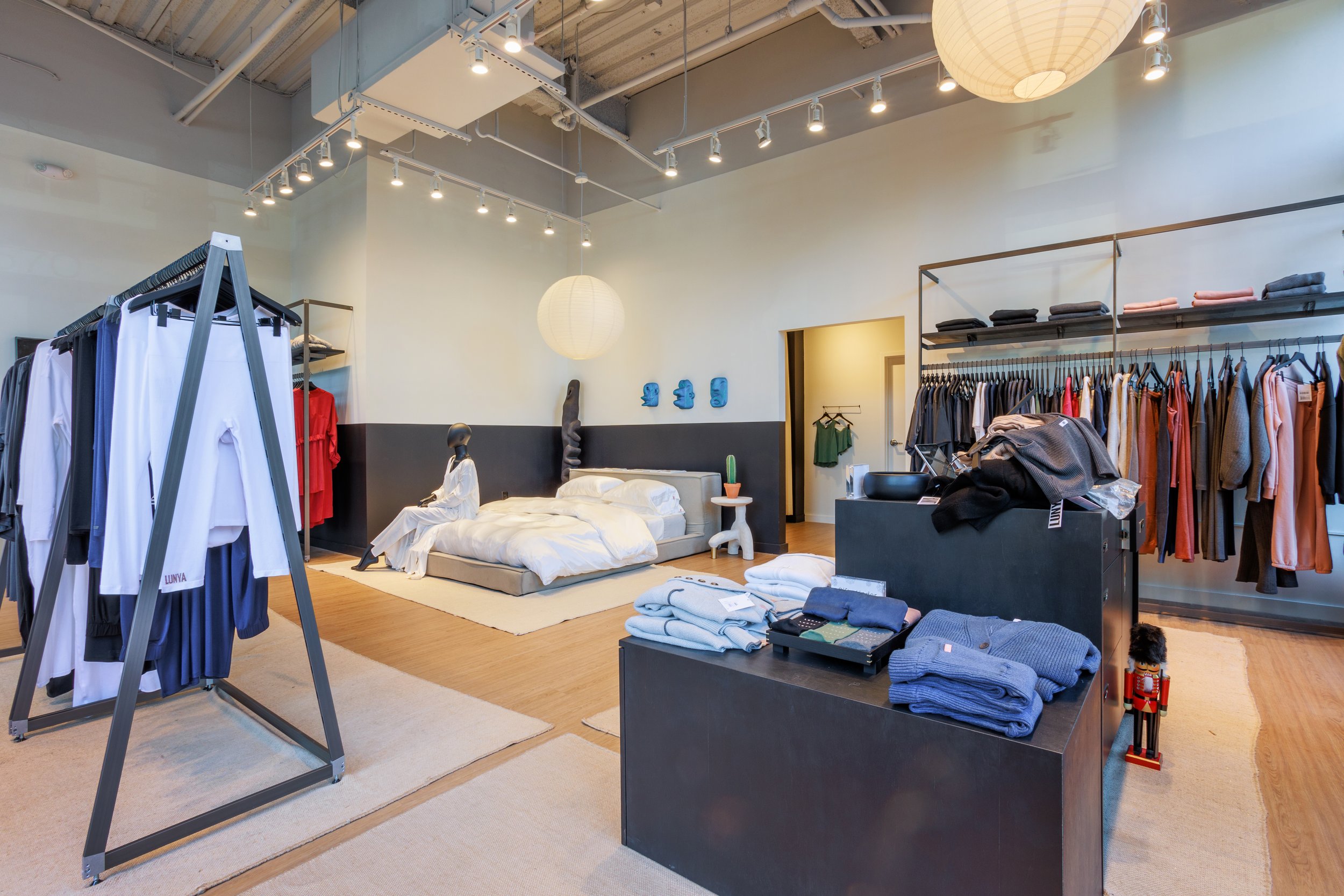 Inside a clothing store displaying racks of clothing, a bed setup with mannequin, and various folded clothes on black fixtures.