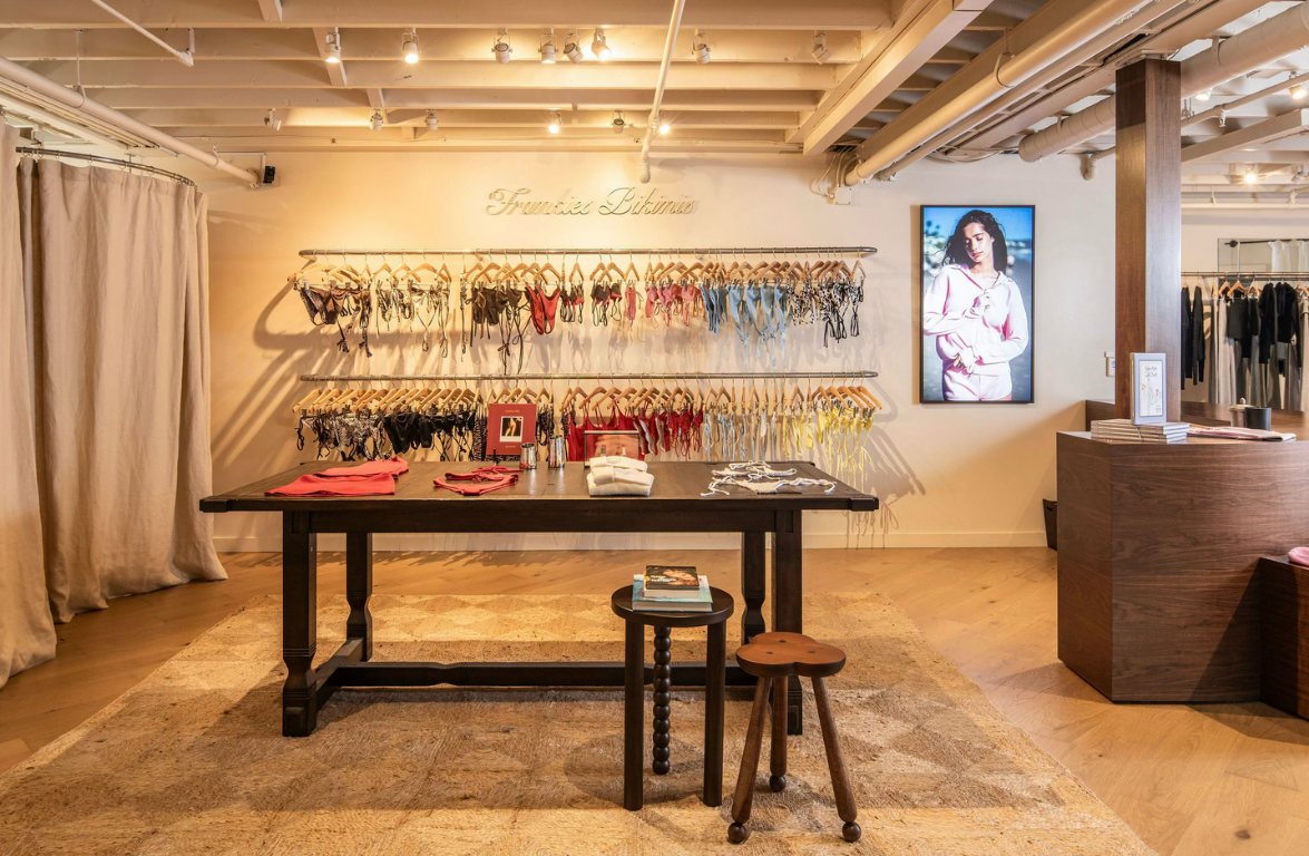 Bathing suit shop interior with racks of bras and panties on the wall, a wooden table with folded clothing and accessories, a digital display of a woman, and a cozy ambiance.