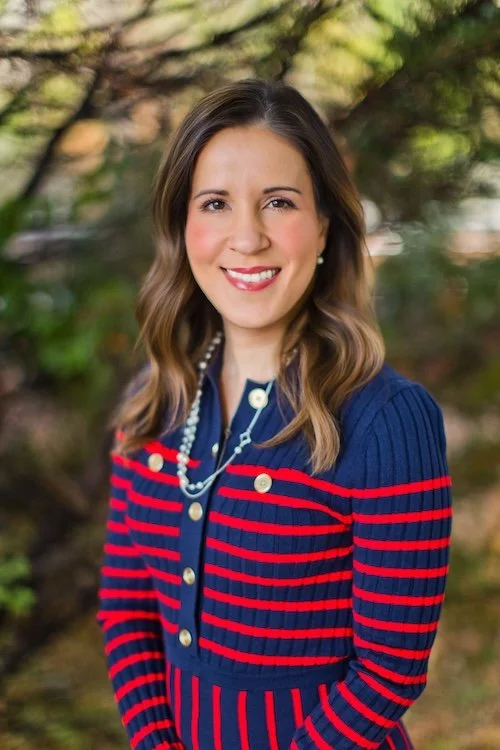 A woman outdoors in front of trees, wearing a navy blue sweater with red stripes and a pearl necklace, smiling at the camera.