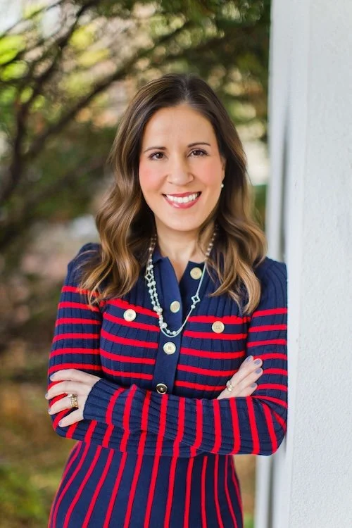 A smiling woman with wavy brown hair, wearing a navy blue jacket with red stripes and gold buttons, standing outdoors beside a white wall with trees in the background.
