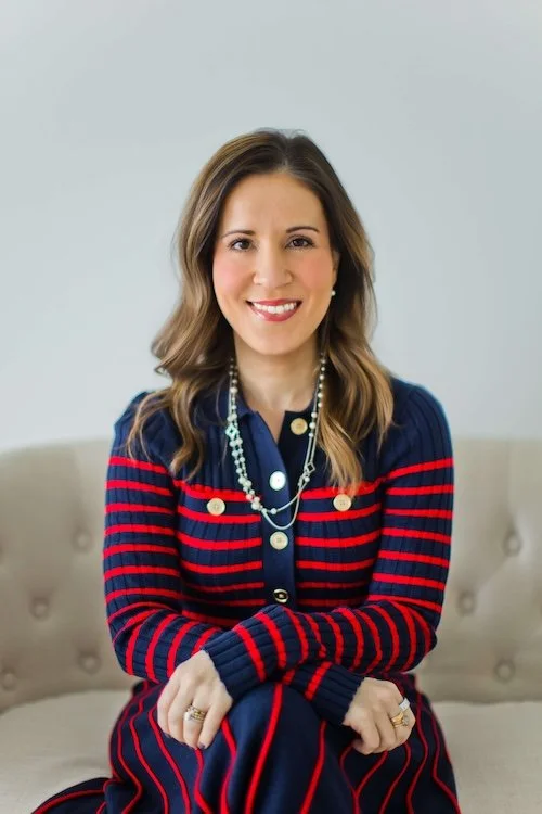 A woman with shoulder-length wavy brown hair, wearing a navy blue and red striped cardigan over a navy blouse, sitting on a beige sofa with a neutral background.