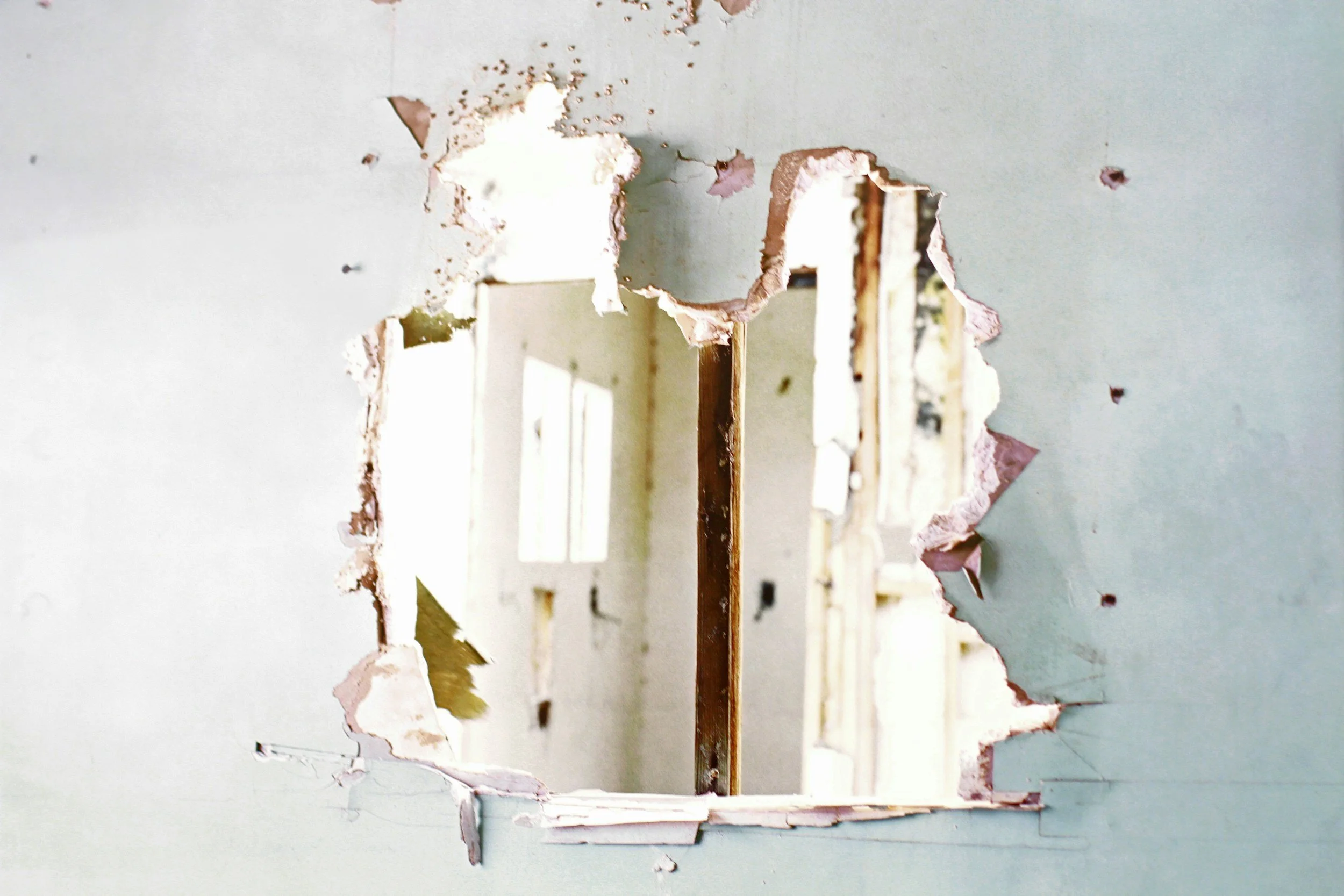A hole in a drywall wall exposing the interior wall studs and framing, with a view into an interior room of a house under renovation.