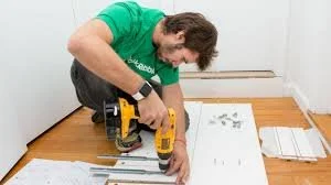 A man installing baseboards using a cordless drill in a room with hardwood floors.
