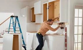 Woman installing kitchen cabinets with a ladder nearby in a partially renovated kitchen.