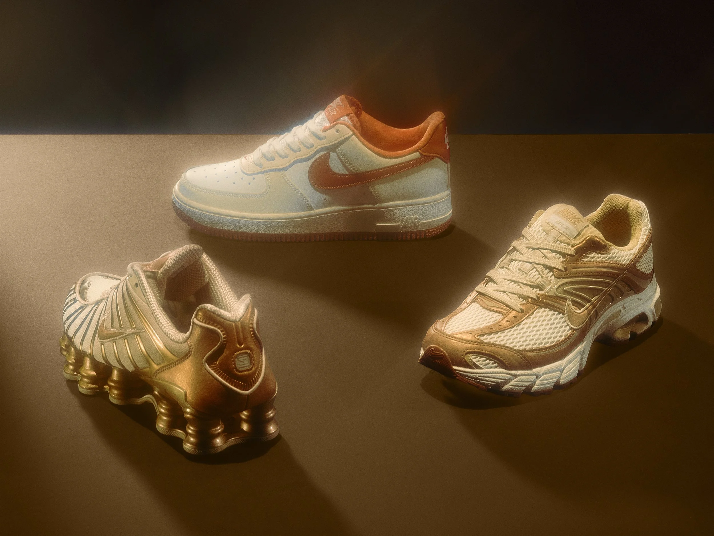 Three pairs of Nike sneakers displayed on a dark surface with a lighting spotlight, including a white and orange pair, a gold pair with a spike sole, and a white and beige pair with a chunky sole.