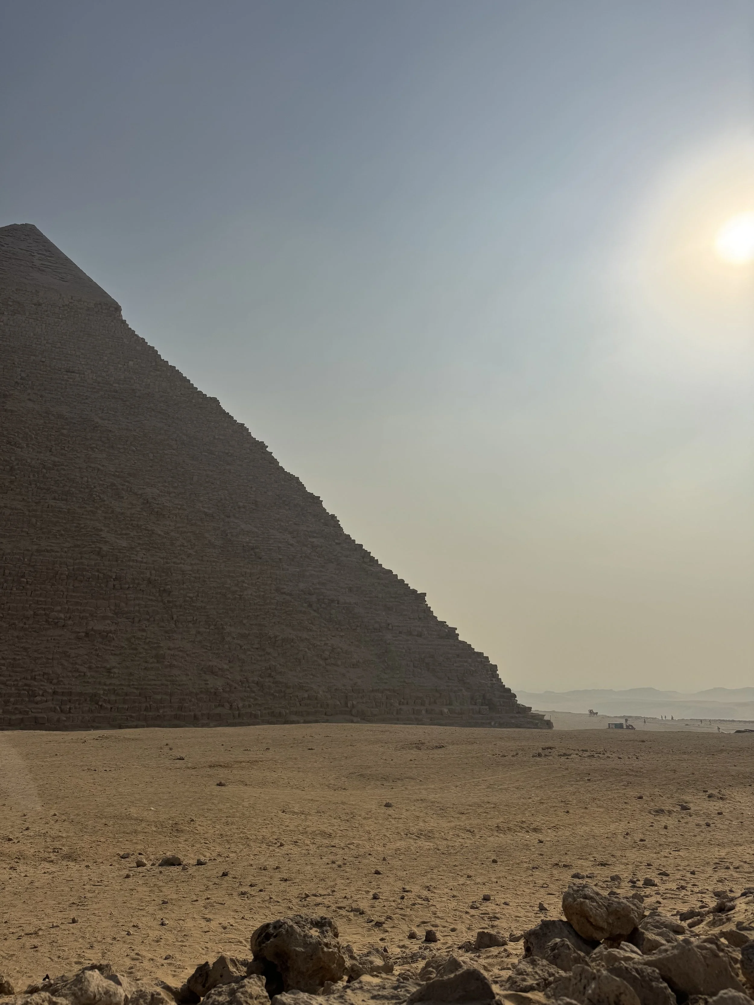 A desert landscape with a large pyramid, sandy ground, and a hazy sky with the sun shining.