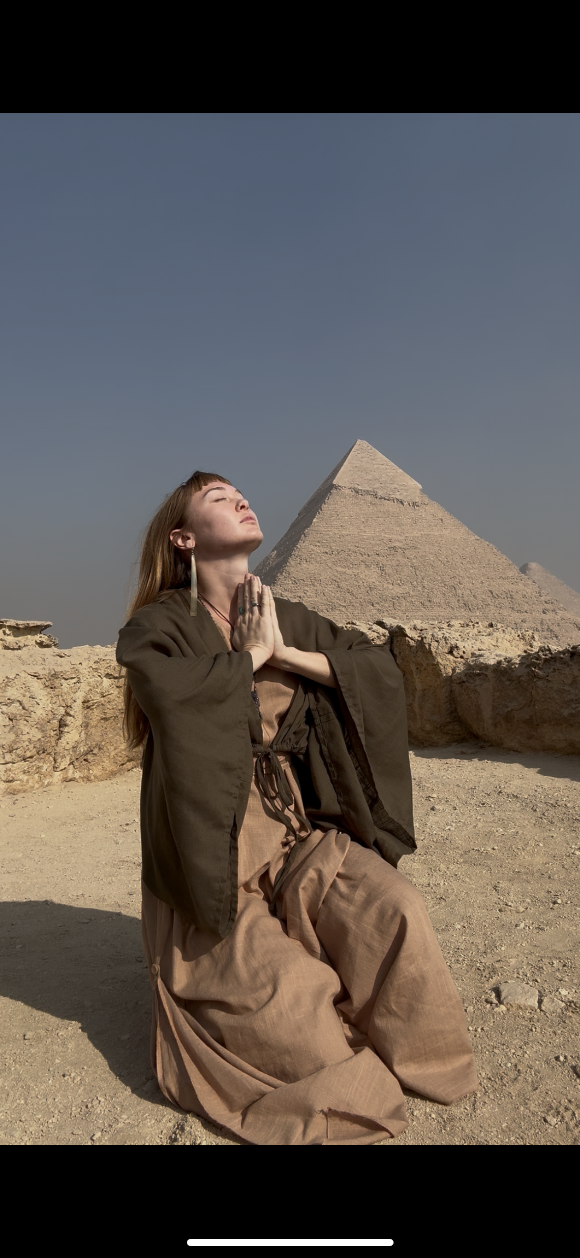 A woman with long brown hair and a brown robe kneels on desert ground with closed eyes and hands pressed together in prayer, in front of the Great Pyramid of Giza under a clear blue sky.