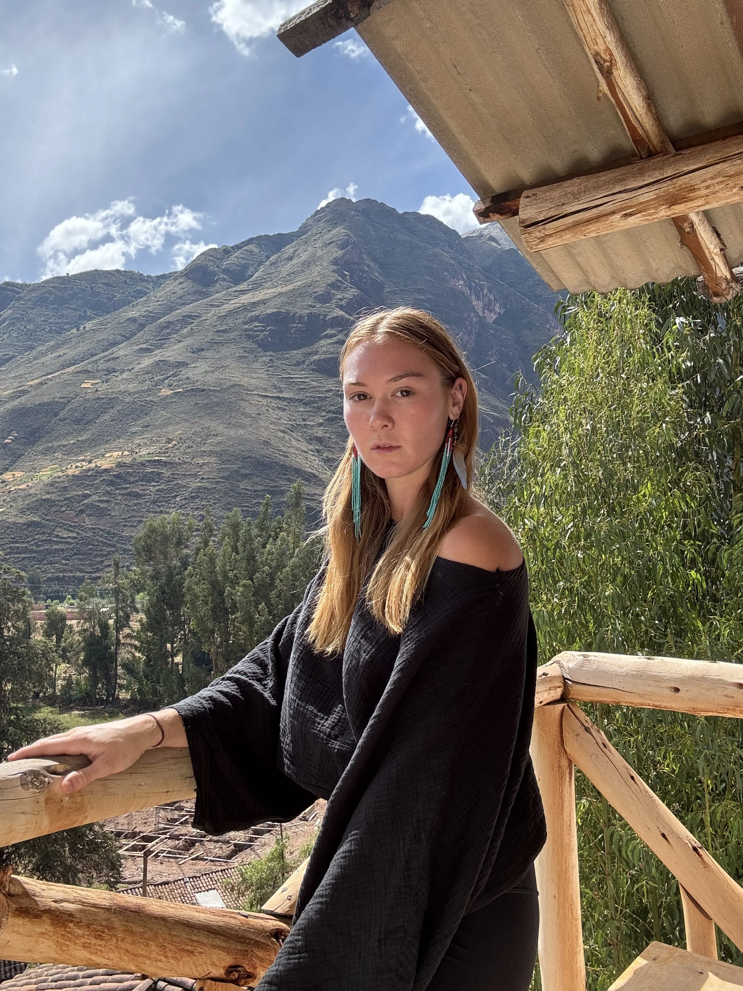 Young woman with long blonde hair wearing a black off-shoulder top and colorful feather earrings, standing on a wooden balcony with a mountain landscape in the background under a partly cloudy sky.