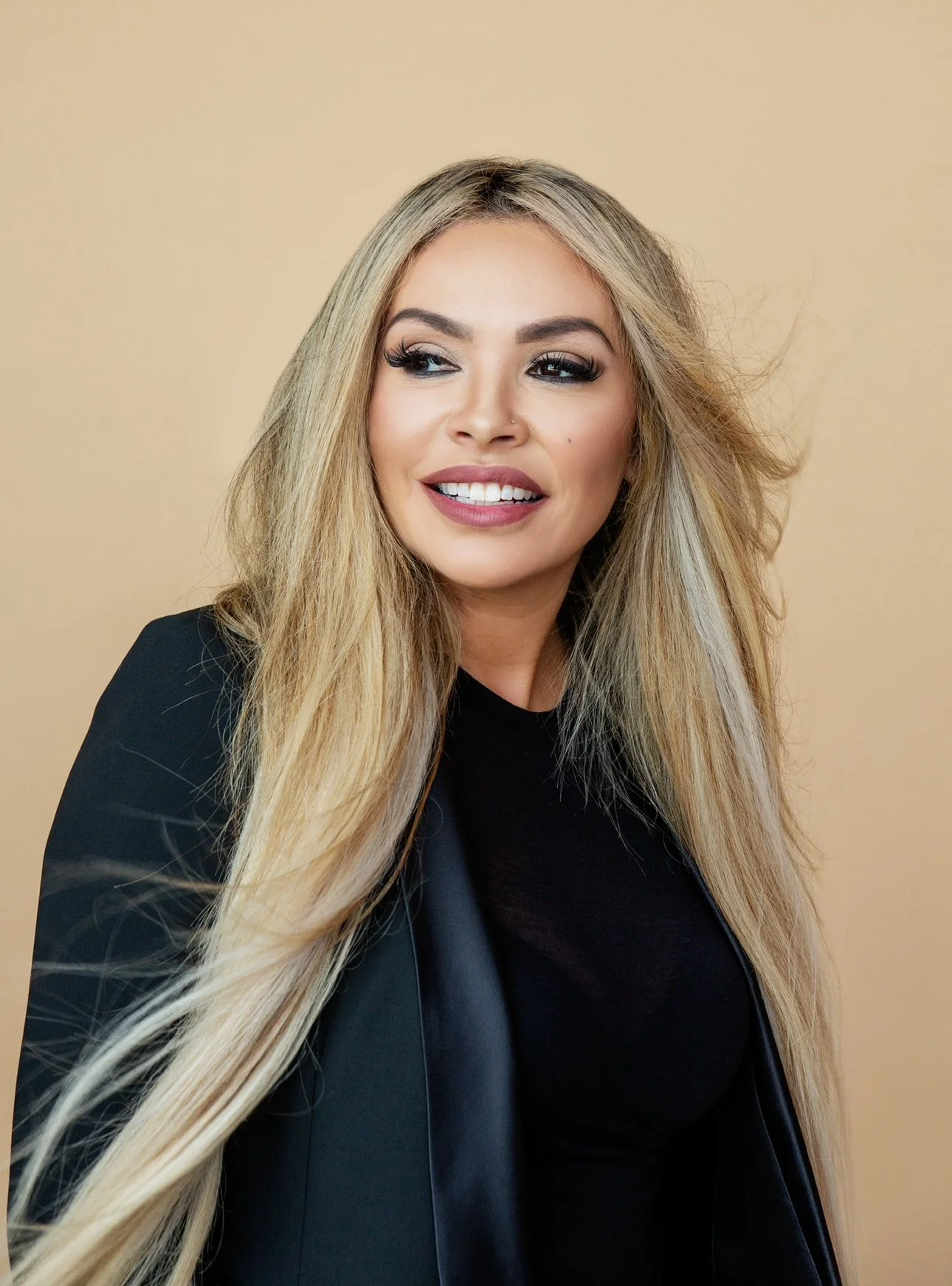A blonde woman with long hair, wearing a black top and blazer, smiling and looking to the side against a beige background.
