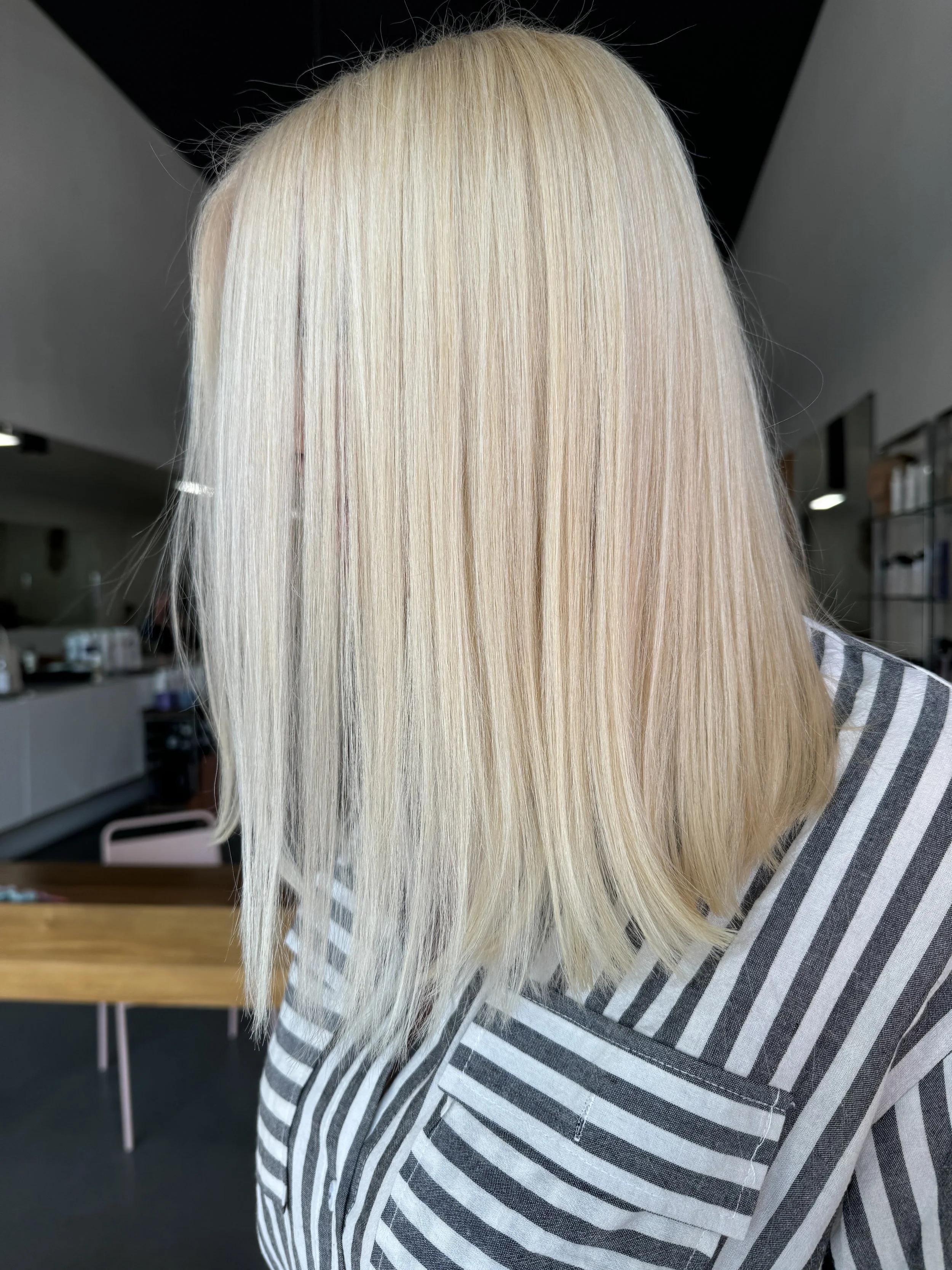 A young woman with platinum blonde shoulder-length hair and light makeup, wearing a light blue and white striped shirt, looking off to the side against a plain white background.