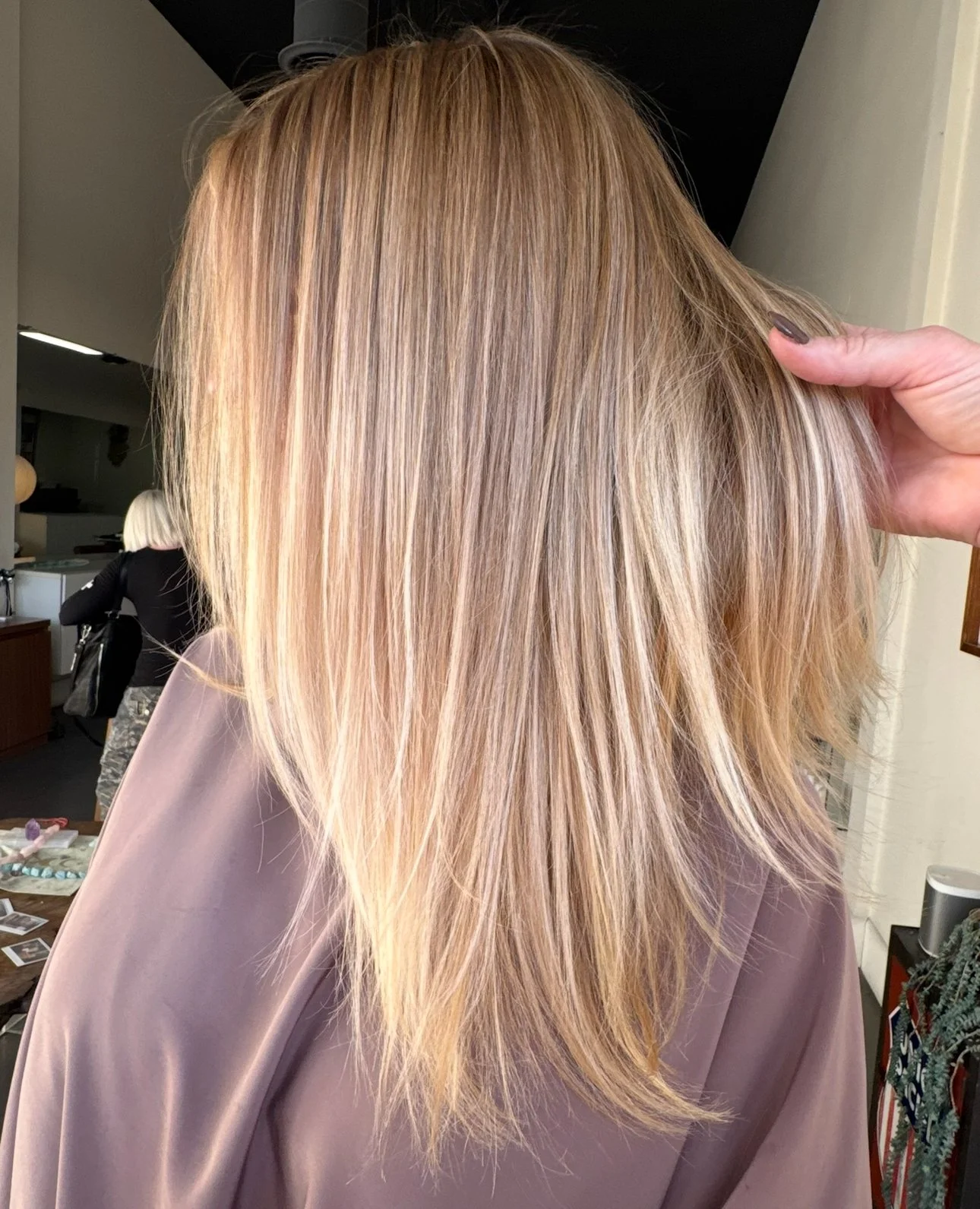 Close-up of a woman with blonde hair in layers, held by a person whose hand is visible, in a hair salon or similar setting.