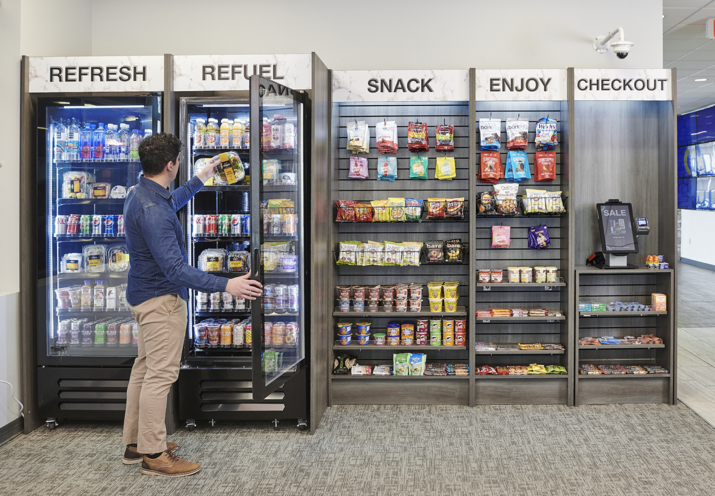 A person in casual clothing is selecting a snack from a refrigerated vending machine in a convenience store, with snack options on display behind him, and a shelf of various snack products labeled 'SNACK' to his right.