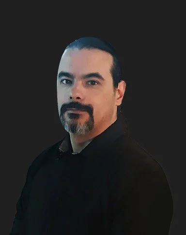 Retrato de un hombre con barba y bigote, con cabello oscuro, usando una camisa negra, con fondo negro.