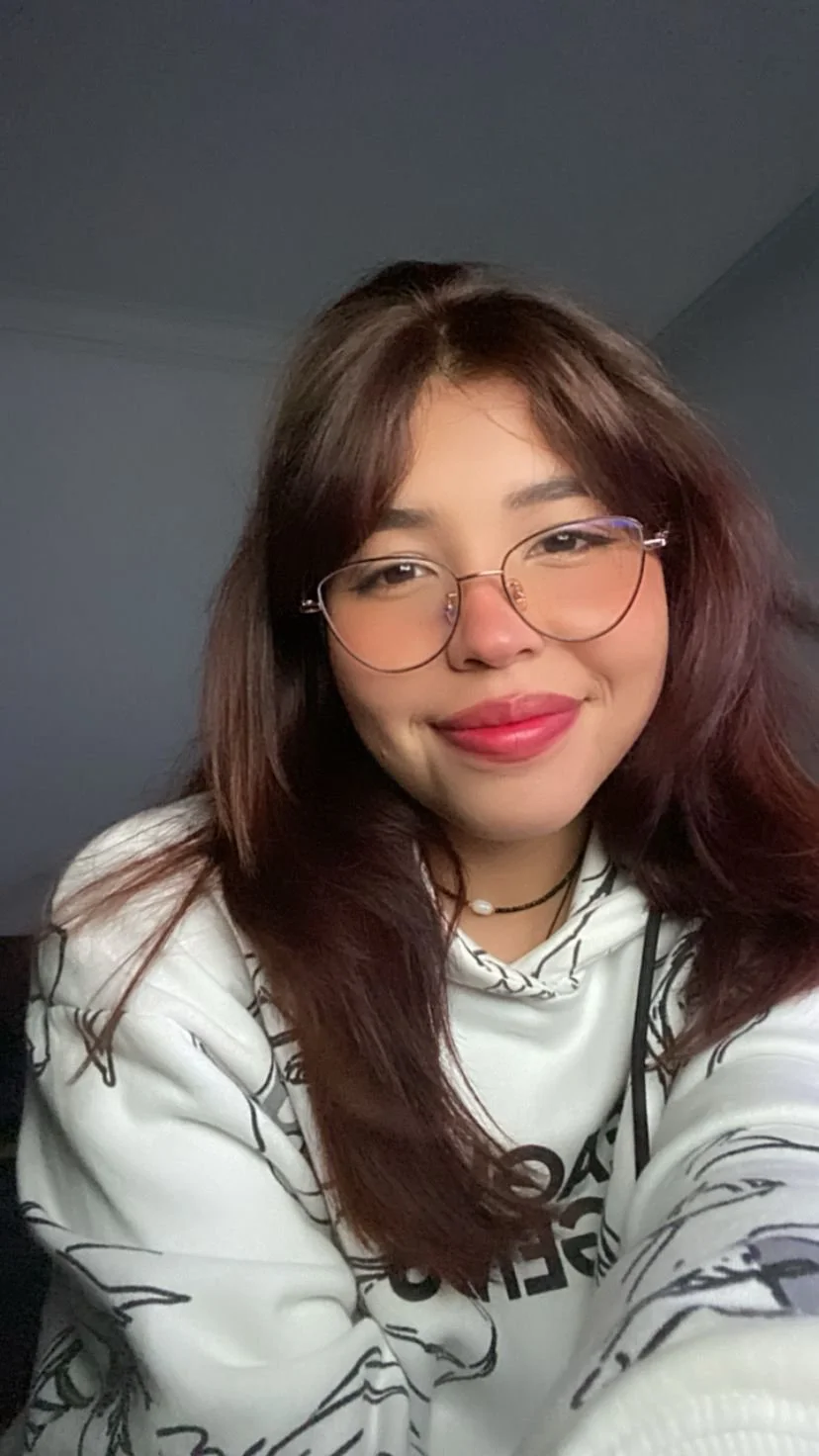 Retrato de una joven mujer de cabello castaño oscuro, gafas redondas y sonrisa sutil, posando en un ambiente interior.