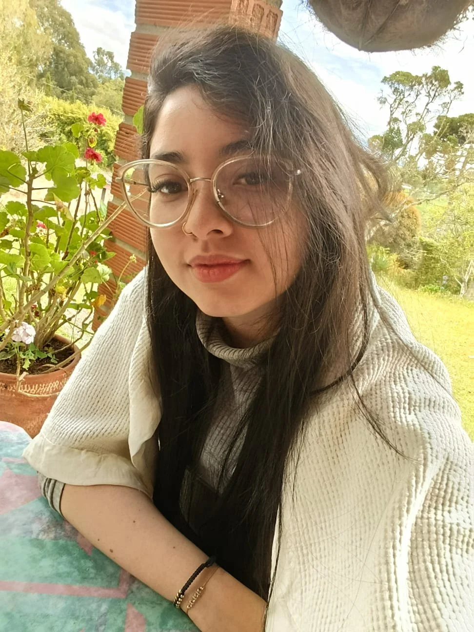 Joven mujer con gafas grandes, cabello largo y oscuro, sonriendo en un entorno exterior con plantas y árboles.