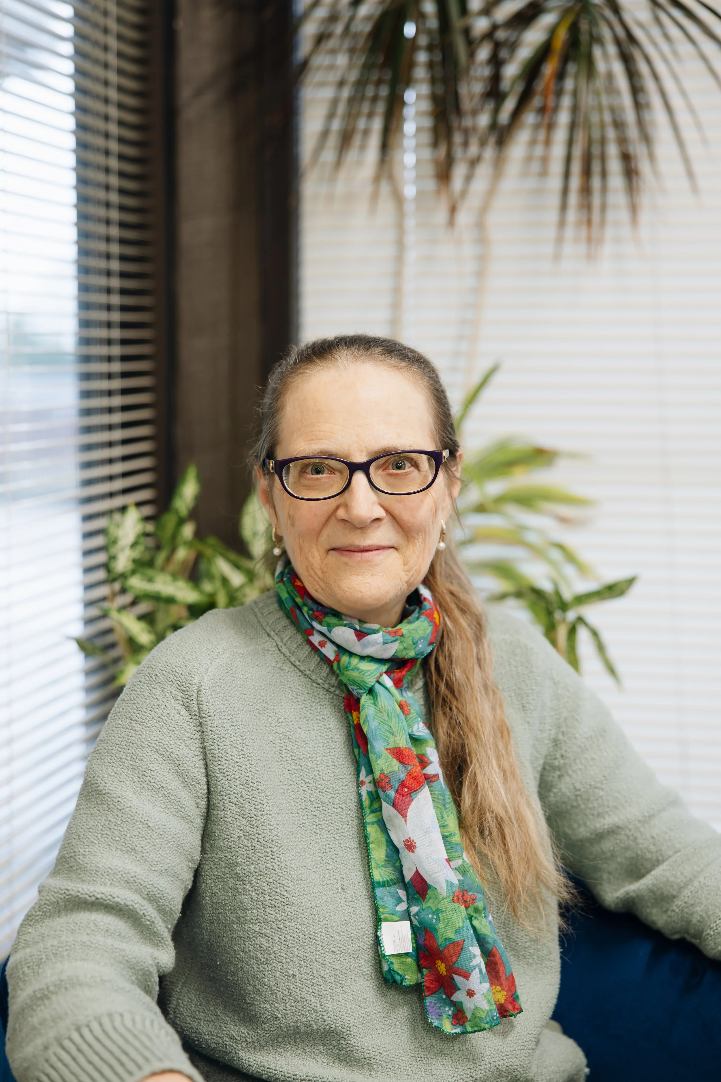 An elderly woman with glasses, wearing a green sweater and a colorful holiday-themed scarf, sitting in front of a window with blinds and green plants in the background.