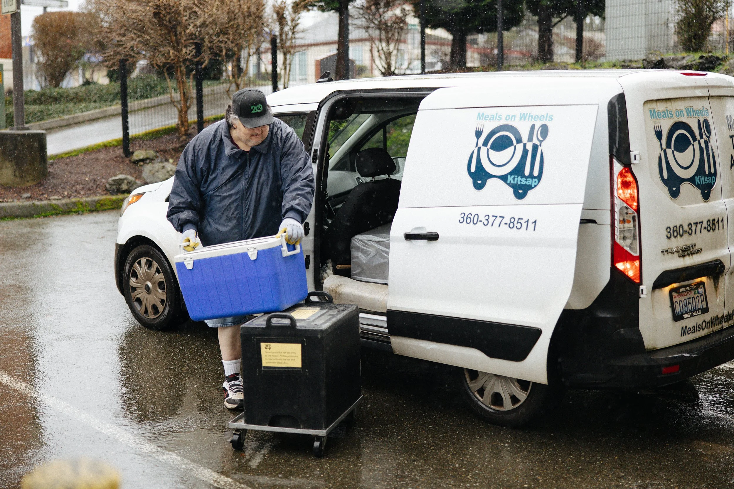 A man loading a cooler into a Meals on Wheels delivery van on a rainy day.