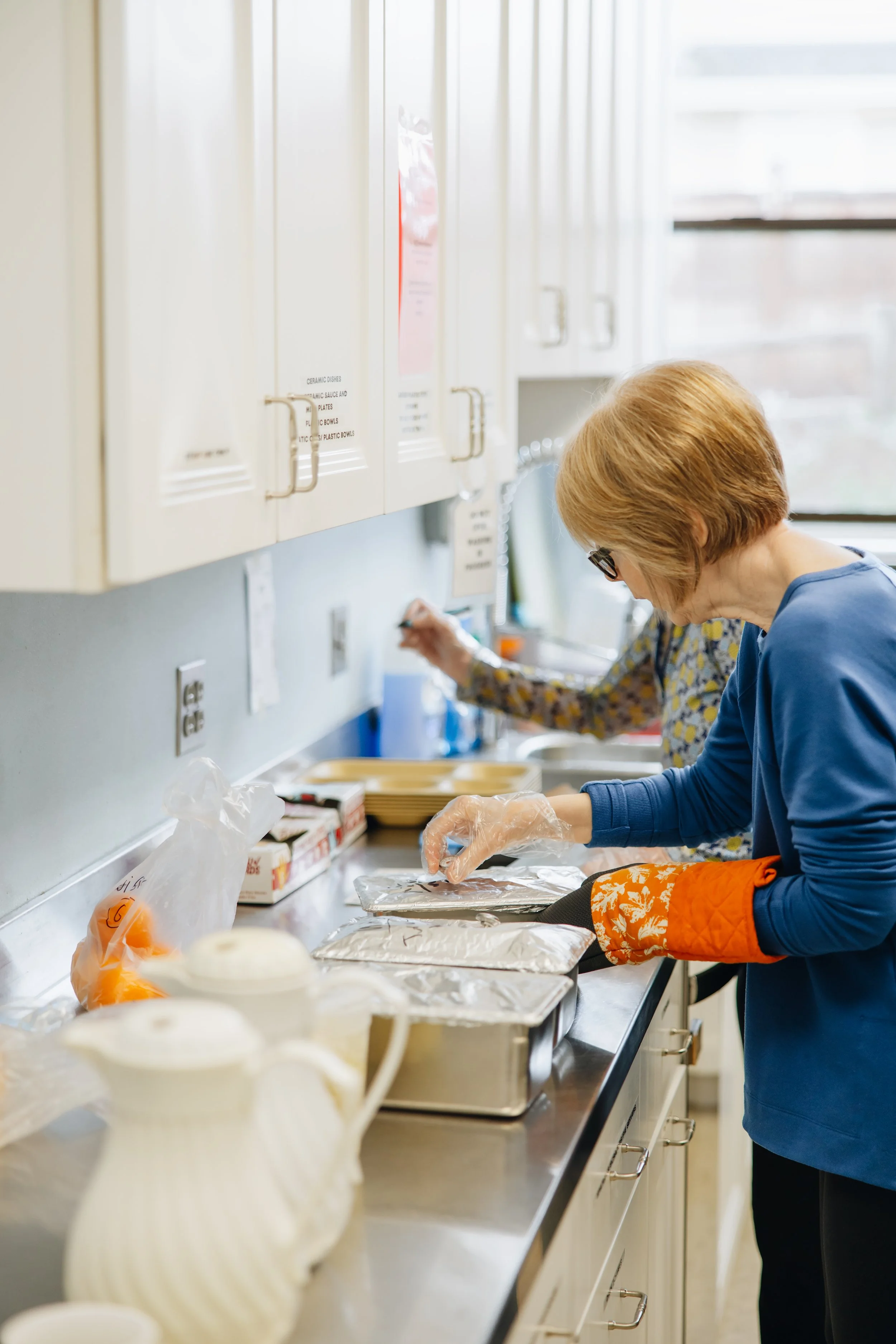 Two women preparing food in a kitchen, wearing gloves and aprons, with foil-wrapped trays on the counter.
