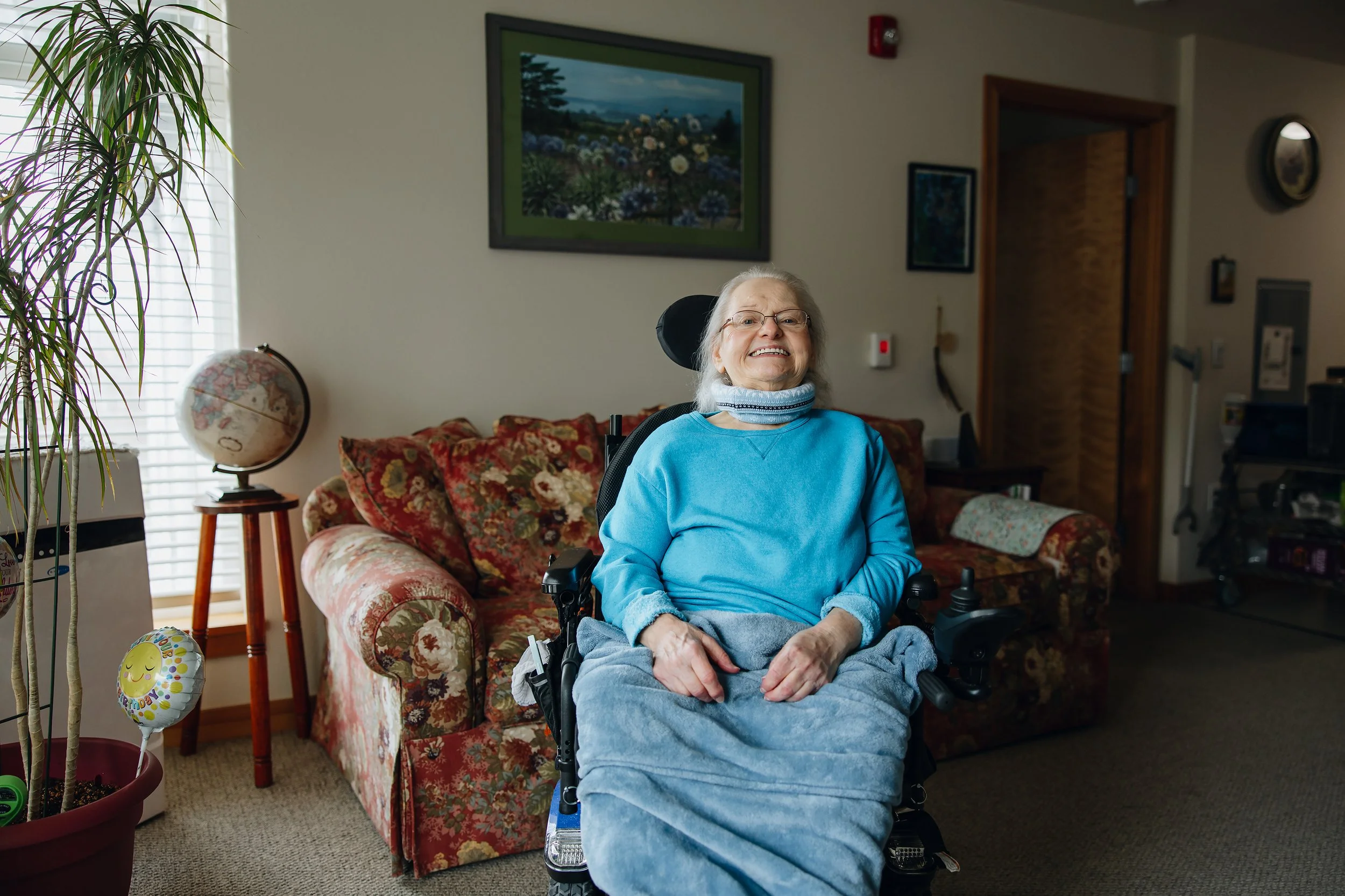 An elderly woman with white hair and glasses smiling while sitting in a wheelchair in a living room. She is wearing a blue sweater and has a gray blanket over her lap. The room features a floral-patterned sofa, a globe on a stand, and a potted plant. There are framed pictures on the wall behind her.