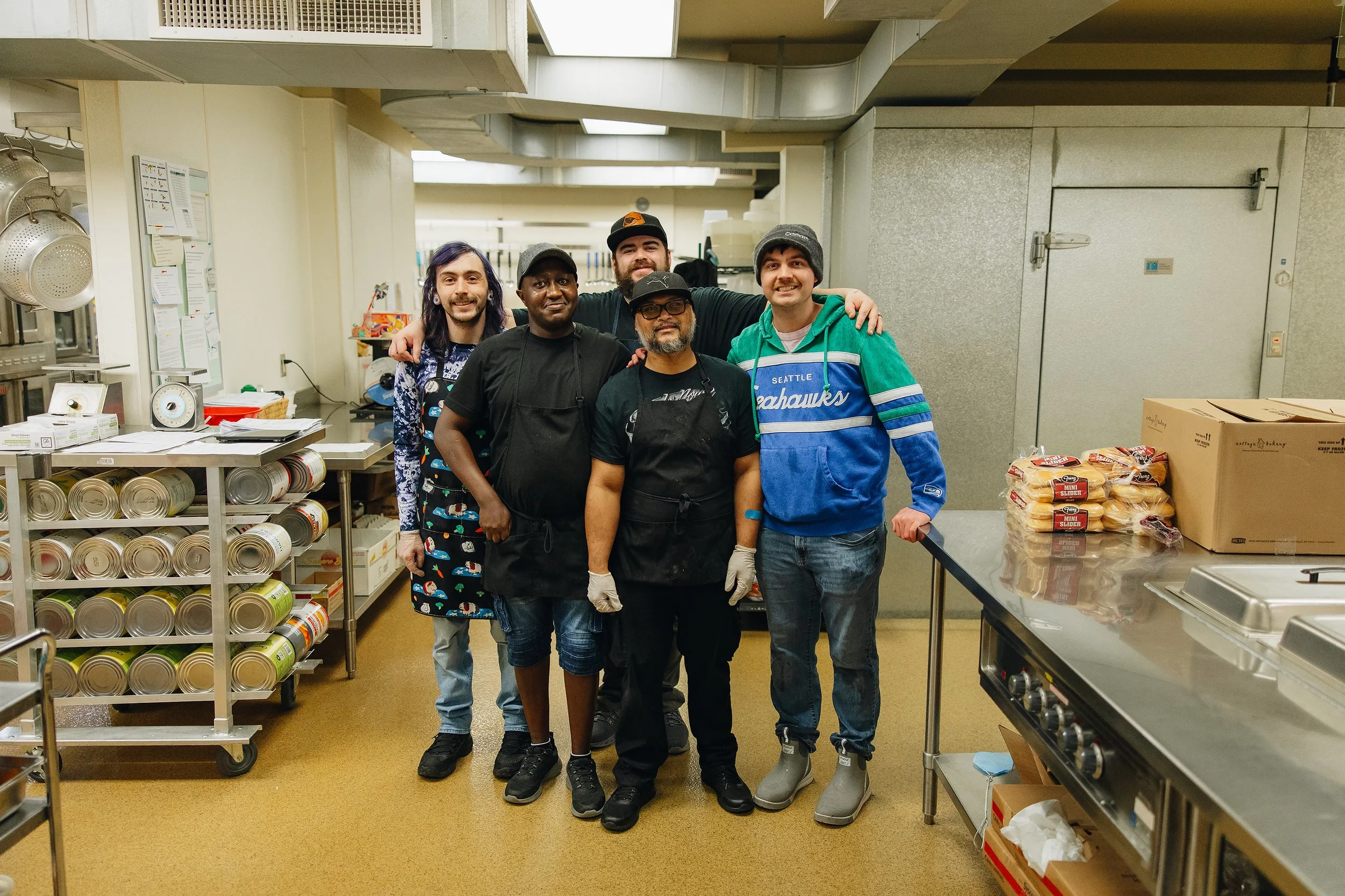 Five people standing together in a kitchen, smiling at the camera. Two are wearing aprons, indicating they are cooks, and the others are casually dressed. The kitchen has shelves with canned goods and boxes, and a counter with packaged food items.