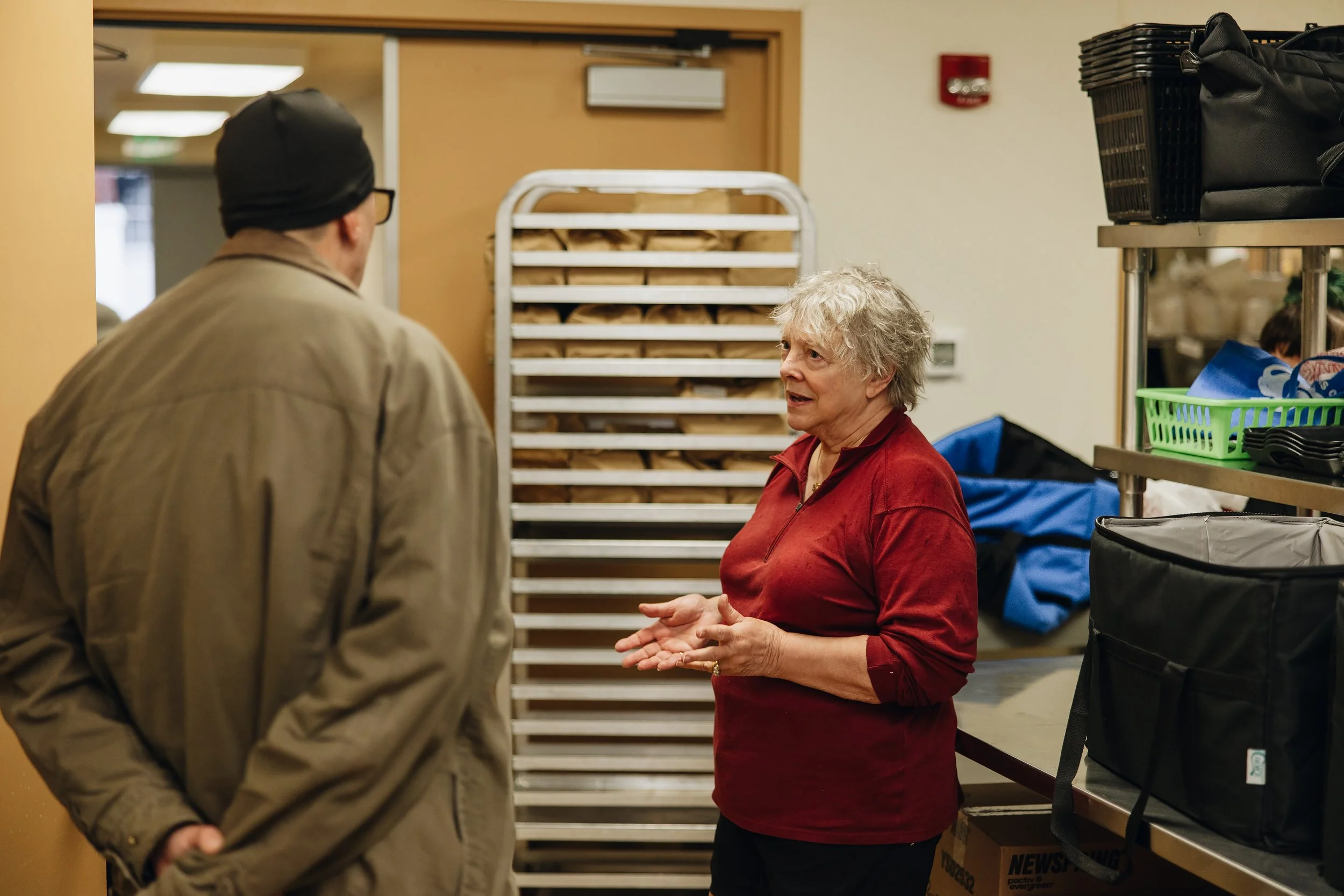 A woman is talking to a man in a food pantry or food bank, with shelves and racks of baked goods and bags around them.