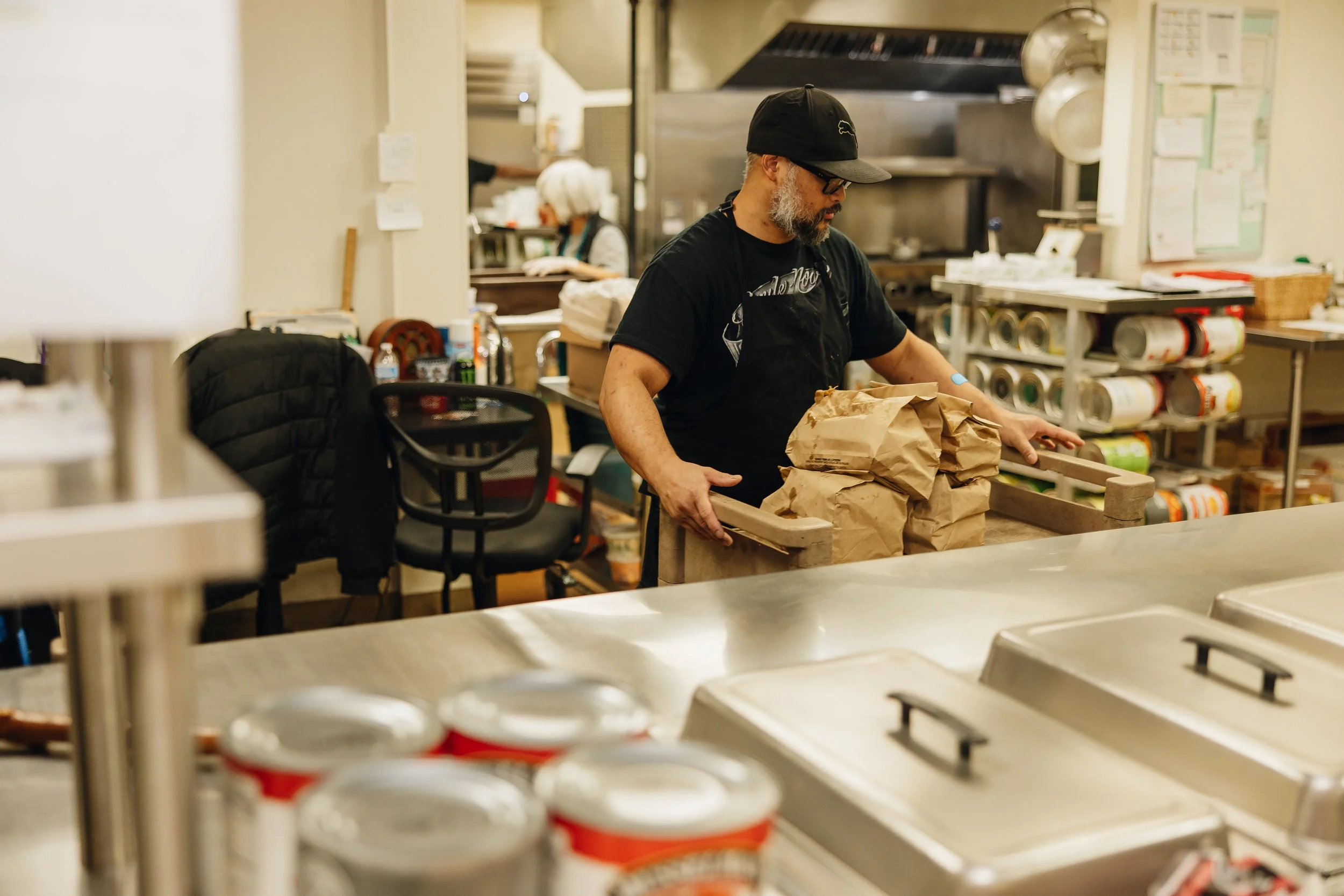 A man wearing a black cap and black apron is packing paper bags in a kitchen or food preparation area. He has a beard and glasses. In the background, there is a woman wearing a white hair cover and a black apron, working at a counter, and shelves with canned goods and other kitchen supplies.