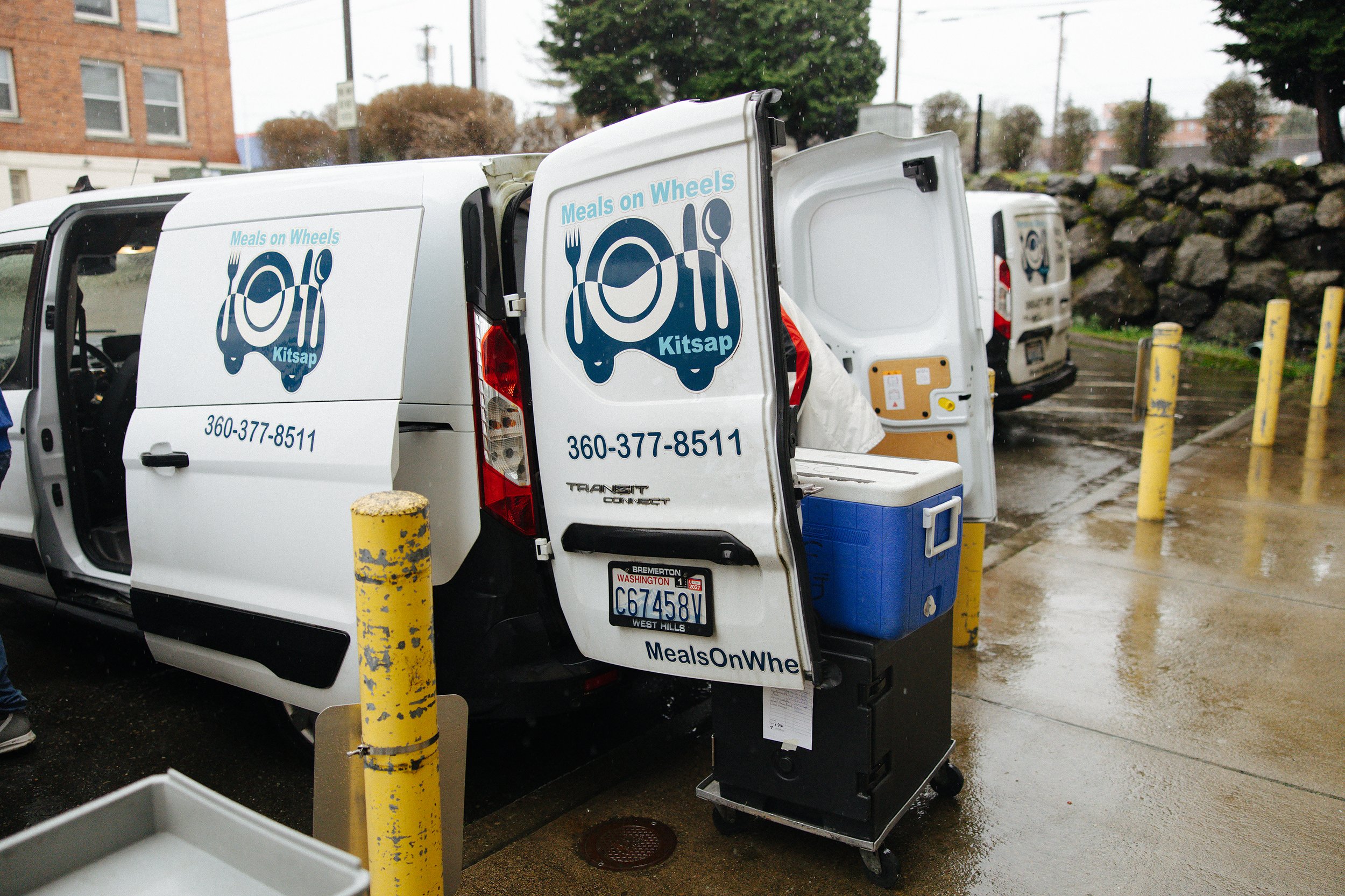 A white delivery van with 'Meals on Wheels Kitsap' logo and contact information parked in a wet parking lot on a rainy day, with a blue cooler and thermoses visible near the open back door.