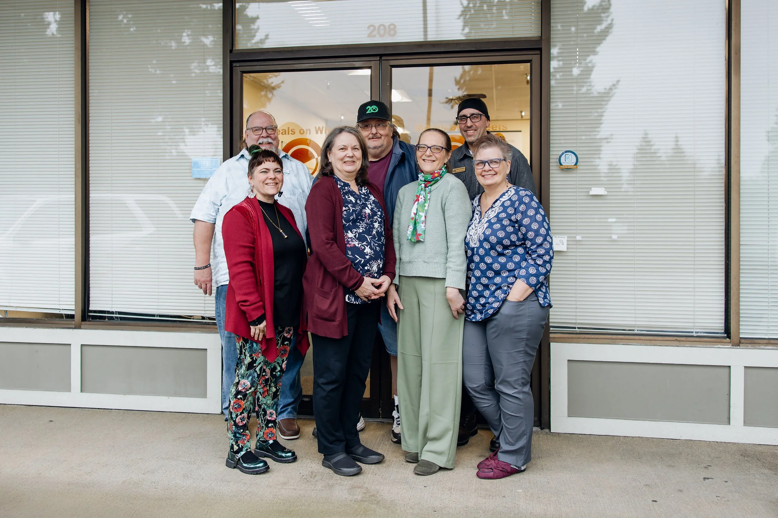 Group of seven people standing outside in front of a building with large windows, smiling for the camera.